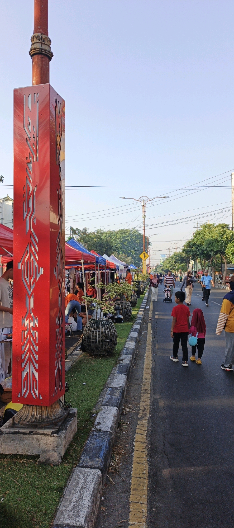 This image depicts a street scene in Sidoarjo, Indonesia, likely during a market or festival event. In the foreground, a large, ornate red pillar with intricate geometric cutouts stands prominently on the left. To its right, a row of market stalls with colorful tents (red, blue, white) are set up along the sidewalk. Several people are browsing or tending to the stalls. Beyond the stalls, a street stretches into the distance, lined with trees on the right side. Various individuals, including adults and children, are walking on the street and sidewalk. Two young children, a boy in a red shirt and a girl in a pink hijab, are walking away from the camera. In the background, streetlights, power lines, and buildings are visible, along with more trees and people. The sky is clear and blue. The overall atmosphere suggests a bustling outdoor event.