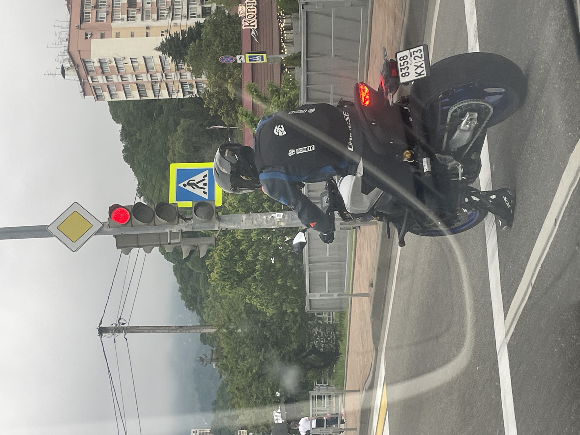A medium shot shows a motorcyclist at a red light in Sochi, Russia. The motorcycle is a sport bike with a blue rim and a license plate that reads "8358 KX 23". The rider is wearing a black motorcycle jacket with "Dainese" and "FCMOTO" logos, gloves, and a helmet. A traffic light shows a red light, with an additional diamond yellow warning sign. A pedestrian crossing sign can also be seen. In the background, there are buildings, trees, and a mountain. A sign above the biker reads, "Koby." The street has white lane markings. Other pedestrians and white vehicles can be seen at a distance.