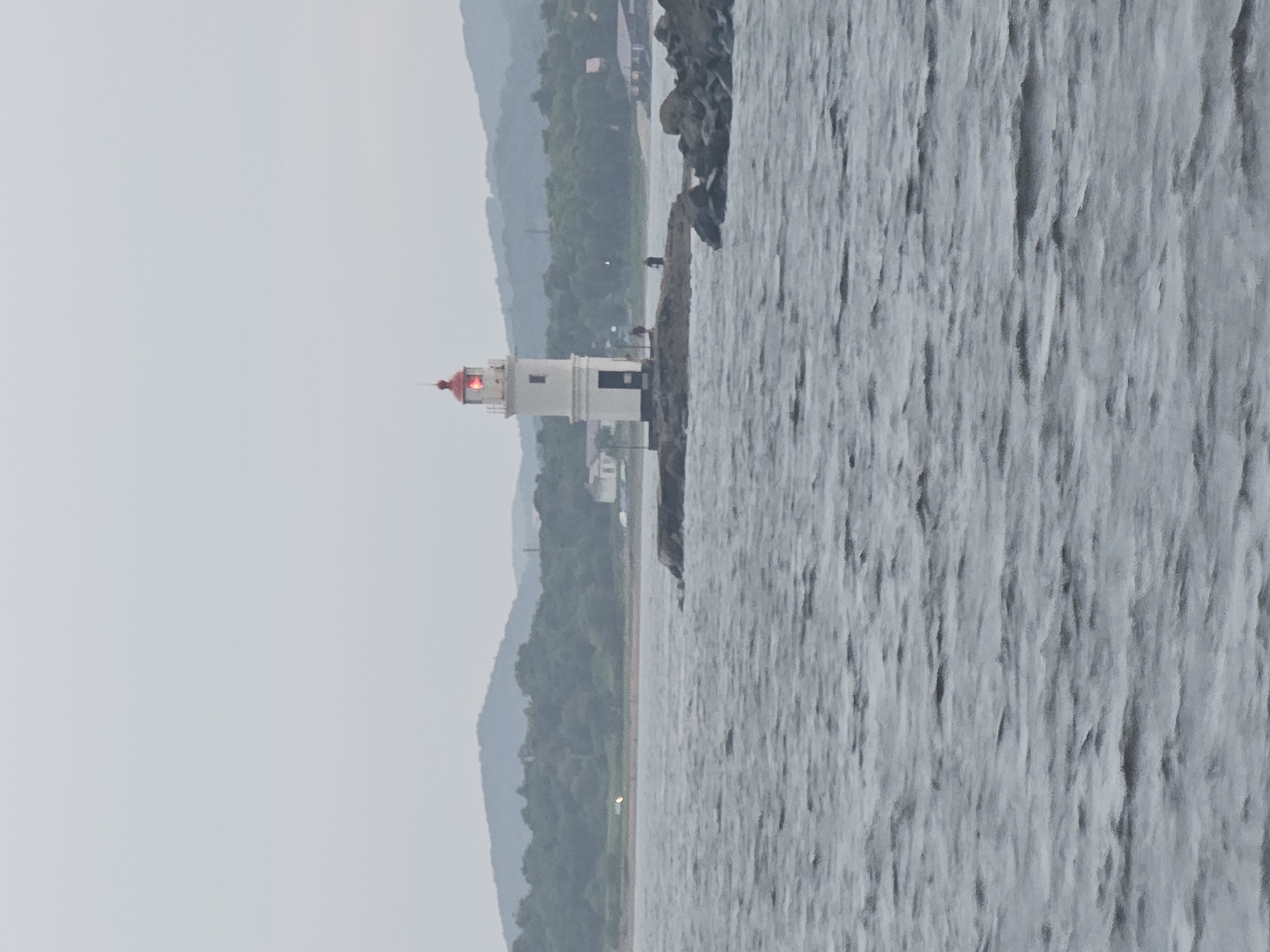 The image shows a white lighthouse with a red top, situated on a rocky outcrop in the water. The lighthouse has a red light on. There are two small figures standing on the rocks near the lighthouse. The water is choppy. In the background, there is a coastline with trees and hills, and some buildings. The sky is overcast. This is the Tokarevsky Lighthouse in Vladivostok, Russia.