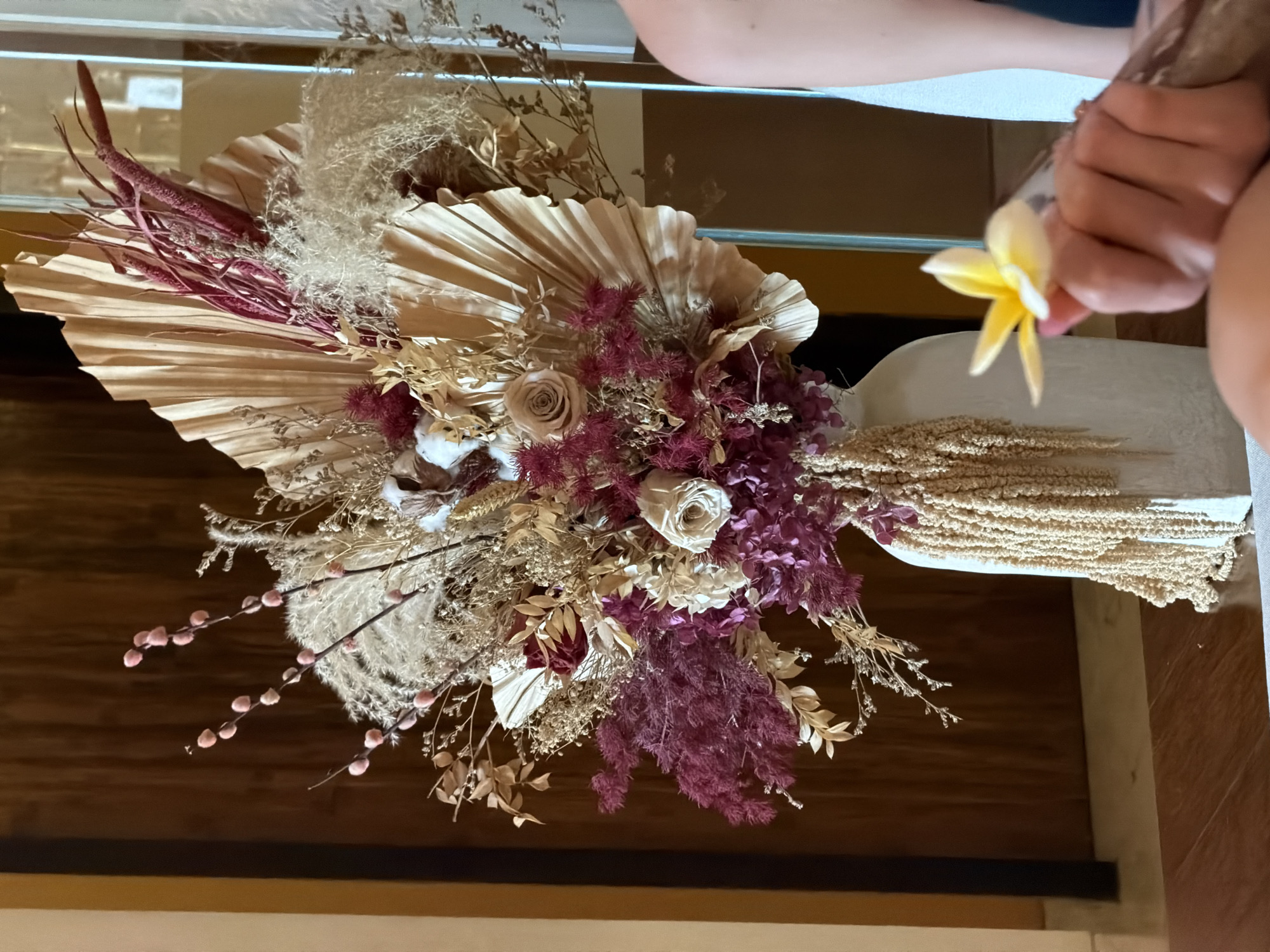 The image displays a detailed view of an elaborate dried floral arrangement situated indoors. The main subject is a large, artistically composed bouquet featuring a variety of preserved plants. Prominently featured are several large, fan-shaped dried palm leaves in a muted beige, forming a natural backdrop. Interspersed within are dried roses in soft, light pink and cream hues, clusters of deep maroon and purple dried flowers, and fluffy, light-colored grasses resembling pampas. Additional elements include elongated maroon seed pods, delicate pinkish-brown catkin-like stems, cotton bolls, and cascading strands of textured beige dried amaranthus or similar plant material, creating a rich tapestry of textures and earthy tones.
On the right side of the frame, a person's hands are visible, gently holding a fresh, vibrant yellow and white frangipani flower. This fresh bloom provides a striking contrast in color and vitality against the subdued palette of the dried arrangement. The arrangement appears to be held in a light-colored, possibly ceramic, vase or container, which is partially visible towards the bottom right.
The background consists of warm, dark brown wooden paneling or furniture, suggesting an interior setting such as a hotel lobby, resort, or a well-decorated home. Ambient lighting illuminates the scene, highlighting the intricate details of the botanical display. There is no visible text in the image. The presence of the frangipani flower strongly indicates a tropical or subtropical location, consistent with North Kuta, Indonesia.