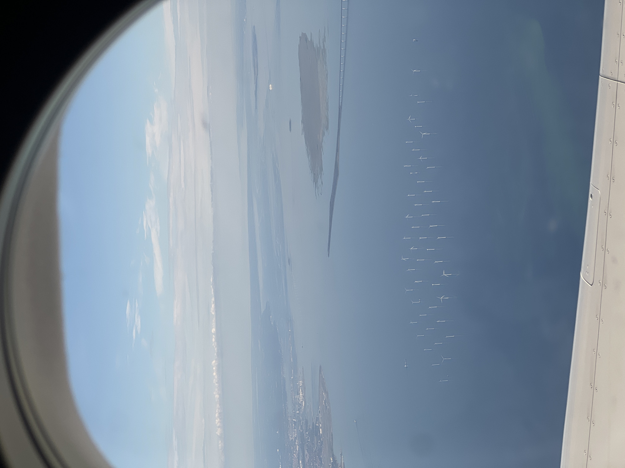An aerial view from an airplane window shows a wide expanse of water, likely the Øresund strait, between Copenhagen and Sweden. In the foreground, a portion of an airplane's wing is visible, with its rivets and metallic surface. The water is a deep blue, dotted with numerous wind turbines, forming an offshore wind farm. These turbines are white, with tall towers and blades extended, standing vertically in the water. In the distance, a long bridge stretches across the water, connecting landmasses. On the left side of the image, a city or town is visible along the coastline, with buildings and infrastructure. The sky above is a pale blue with wispy white clouds, and a hazy horizon. A few ships and boats are also visible on the water. The scene captures a coastal landscape with renewable energy infrastructure and urban development. The location is identified as Kastrup, Denmark, suggesting this view is taken from a flight approaching or departing from Copenhagen Airport.
