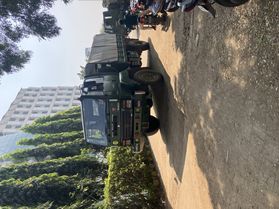 A green Indian Army truck with "NDA" on its bumper sits on a dirt and concrete road, shadowed by the sun. The truck is parked next to a row of green trees and bushes. Behind the truck and trees is a modern building with many windows. Several motorbikes are parked along the side of the road ahead of the truck. Another green truck is visible in the distance.