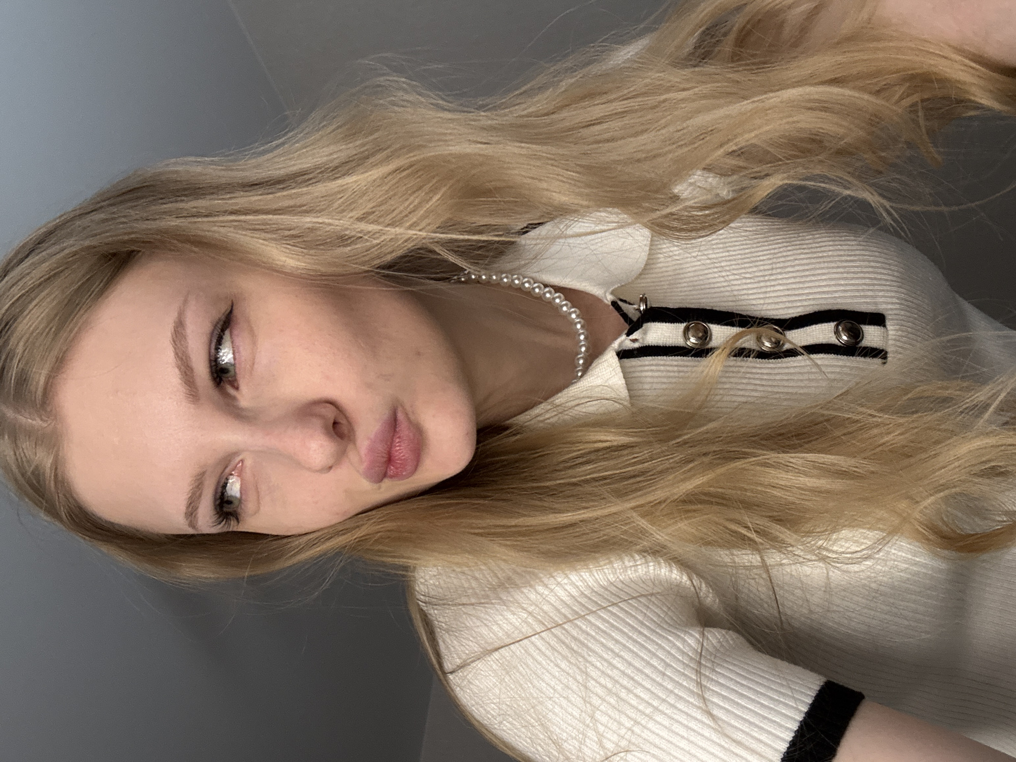 A young adult female is depicted in a close-up, chest-up indoor shot. She has fair skin, long wavy blonde hair, and light-colored eyes. Her head is tilted slightly to her right, and she is looking towards the viewer's left. Her lips are puckered into a "duck face" expression. She wears a white, short-sleeved, ribbed polo shirt featuring a collar and a placket with a vertical black stripe and three round, silver-toned buttons. The sleeves have black trim at the cuffs. A single strand of small, white pearls is visible around her neck. The background consists of a plain, unadorned, muted gray wall, which offers no distinct architectural or environmental features to identify a specific geographic location, including the city of Perm, Russia. The lighting is soft and even.