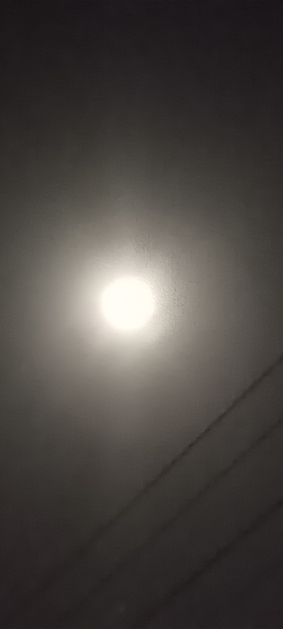 The image shows a bright, circular light source, likely the moon, centered in a gray sky. Diagonal, dark lines, possibly power lines, are visible in the lower right corner. The image is dark and lacks detail, making it difficult to provide further forensic analysis. There is no evidence to suggest that this image was taken in Meuse, Indonesia. No people or interactions are visible.