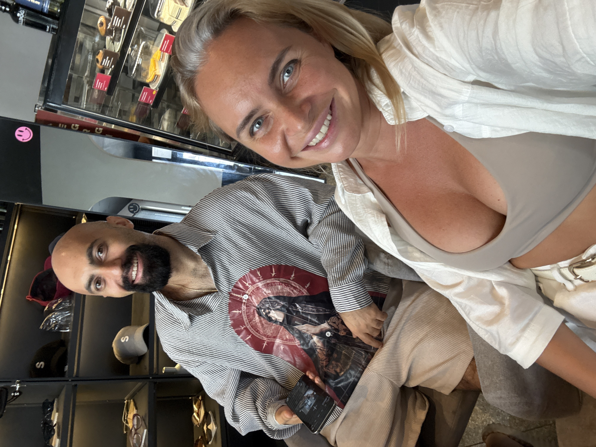 A man and a woman are seated together, posing for a selfie in what appears to be a cafe or a shop selling accessories. The man, with a shaved head and beard, wears a striped shirt with a graphic print. He is holding a phone in his left hand. The woman, with blonde hair, smiles broadly at the camera. She is wearing a light-colored shirt over a top. Behind them, shelves display various items, possibly including hats and sunglasses. In the background, a refrigerated display case filled with cakes and pastries suggests a food establishment. The overall environment appears to be indoors. The presence of cafe-like elements, such as the display case with desserts, suggests a commercial establishment. The context of an accessory shop and a cafe could indicate a boutique or a cafe with merchandise. The setting appears to be a modern, well-lit commercial space.