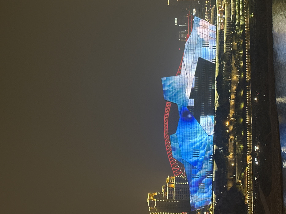 The image depicts a nighttime cityscape of Yuzhong District, Chongqing, China, dominated by the Chongqing Grand Theatre. The theatre, a large, angular, multi-faceted architectural structure, is prominently featured in the middle ground, its glass facade illuminated by a dynamic blue and white light projection, creating patterns resembling flowing water or abstract forms. Behind the theatre, a red arch truss bridge, identifiable as the Dongshuimen Bridge, spans a gap. To the left of the theatre, several high-rise buildings are visible, illuminated with warm yellow lights on their upper sections. The right background shows distant city lights and the faint outlines of additional urban structures. In the foreground, a dark body of water, likely the Yangtze River, reflects subtle light. Above the river, a terraced embankment or park area is visible, illuminated by numerous warm yellow lights highlighting trees and pathways leading up to the theatre structure. No discernible individuals, specific actions, or interactions are visible. The sky is dark, indicating night conditions.
