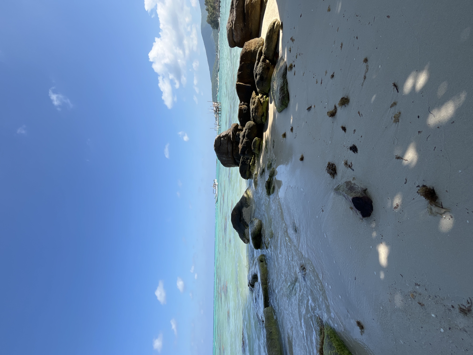 This image depicts a beach scene in Legonipah, Indonesia. The foreground features a sandy beach with scattered rocks and seaweed. The tide is low, revealing wet sand and some submerged rocks. In the middle ground, the clear, turquoise water laps gently against the shore. Several boats are visible in the distance, including a small white vessel closer to the shore and a larger boat with a cabin further out. A wooden pier or structure extends into the water in the middle distance. The background shows a lush green hillside with palm trees and mountains under a bright blue sky with scattered white clouds. The overall environment is serene and tropical. There are no people visible in the image.