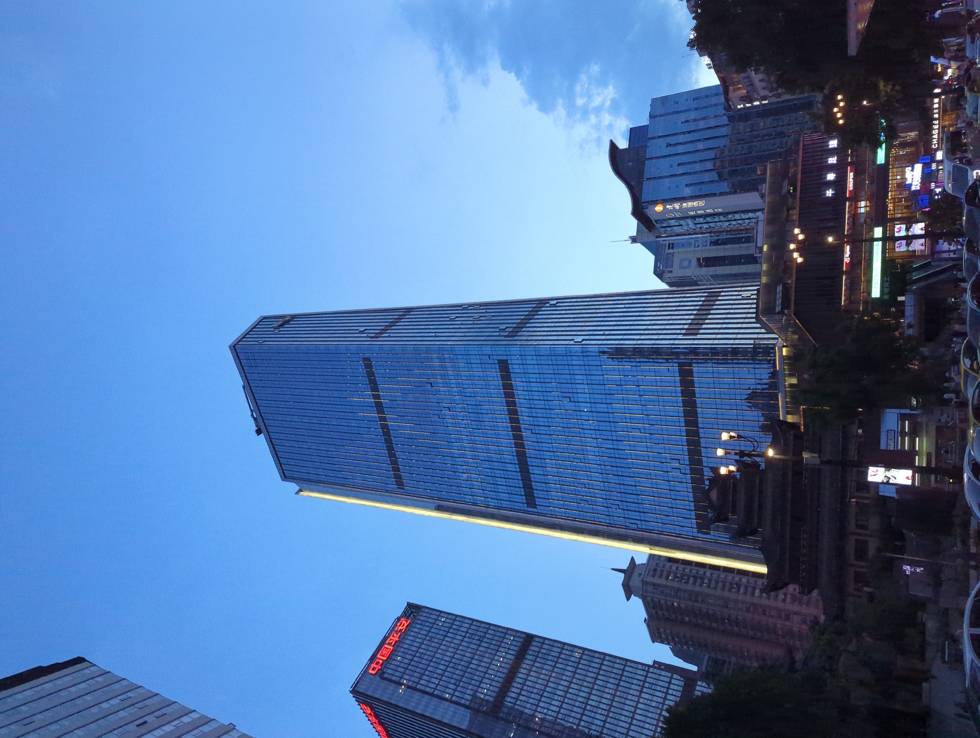 The image depicts a cityscape at dusk in Yuzhong District, China, dominated by modern skyscrapers. The sky is a deep blue, transitioning into twilight. A prominent glass-clad skyscraper with a vertical yellow illuminated strip stands in the center, reflecting the twilight sky. To its left, another tall building displays red Chinese characters that read "中国平安" (China Ping An).
Below the skyscrapers, traditional Chinese architecture is visible, including a multi-tiered dark-tiled roof structure with ornate eaves, illuminated by strategically placed lights. This structure appears to be a temple or cultural building. In front of these buildings, there are streetlights with multiple bulbs.
Further down, a bustling street scene unfolds. Several cars, including a white van and a sedan, are parked or moving on the road. Pedestrians are visible on the sidewalks, and various shop signs with bright neon lights are illuminated, indicating commercial activity. One sign prominently displays "CHASE 环球金融". The overall impression is a blend of modern urban development and preserved cultural heritage.