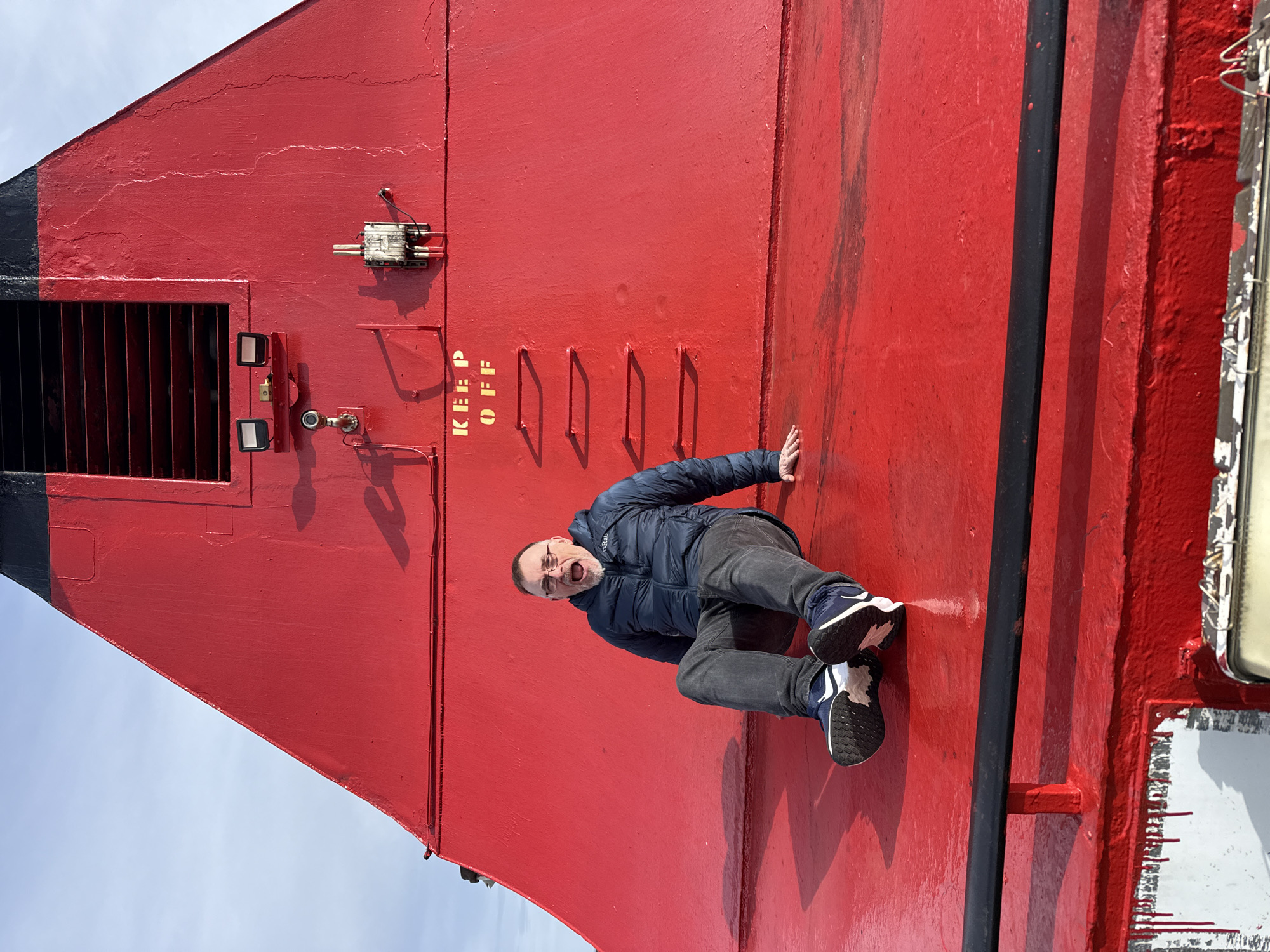 A man in a blue jacket and gray pants is seen seemingly "climbing" a red, large structure. The structure has the words "KEEP OFF" painted in yellow, and it features metal rungs. The sky is visible in the background. The location is Unknown, Unknown.