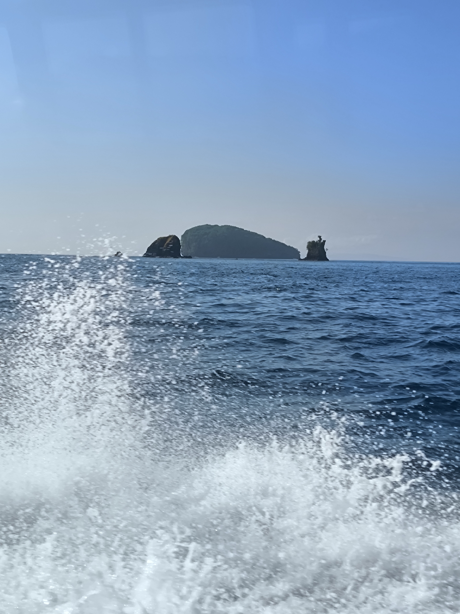 The image shows a body of water with an island in the background. The water is a deep blue color and there are white splashes in the foreground, likely caused by a boat moving through the water. The island is green with rock formations on either side. The sky is a clear, light blue. The location is unknown.