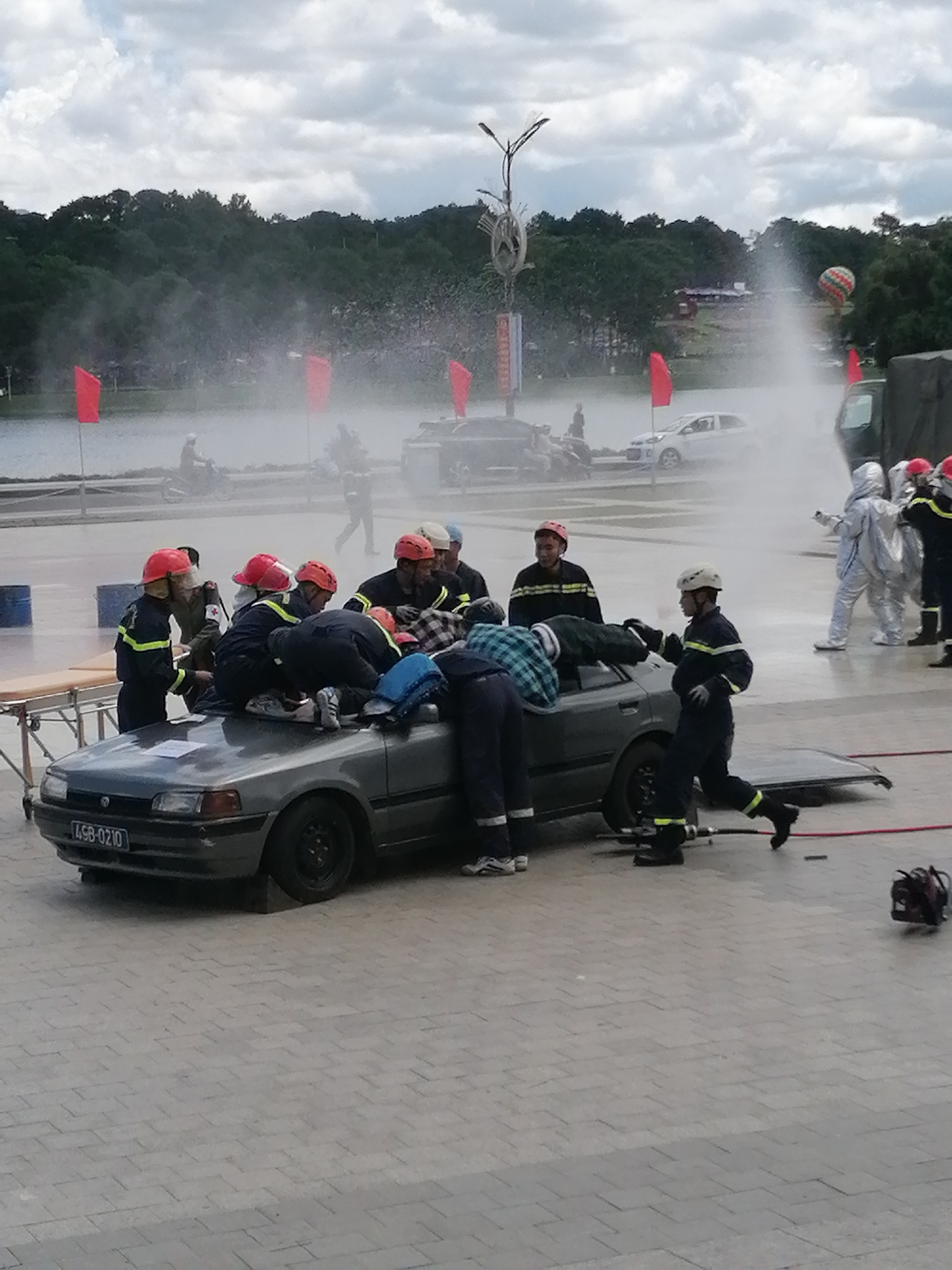 This image captures a simulated rescue operation in Da Lat, Vietnam. A group of individuals, appearing to be firefighters in uniform with helmets, are gathered around a dark-colored sedan. The car is positioned on a paved surface, with its hood open and doors ajar, suggesting a scenario involving a vehicle accident or entrapment. Some of the individuals are leaning into the car, seemingly attending to someone inside or performing extrication maneuvers. Others are standing nearby, observing or assisting. In the background, a body of water can be seen, possibly a lake or river, with trees and vegetation lining its edge. A few red flags are planted along the waterfront, and a hot air balloon is visible in the distance. Water sprays are also present in the background, suggesting a controlled demonstration or drill. The overall scene implies an emergency response training exercise, showcasing the efforts of rescue personnel.