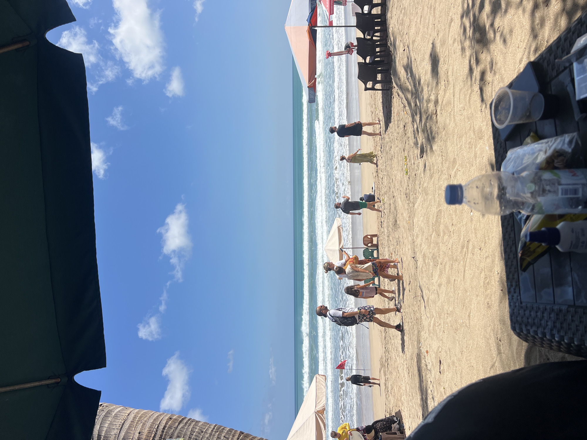 The image captures a lively scene on Kuta Beach in Bali, Indonesia. The bright sun illuminates the sandy shore where numerous people are enjoying a day at the ocean. In the foreground, a table is set with a water bottle, an empty cup, and a plastic bag, suggesting a relaxed setting under a shade umbrella.
Further out on the beach, groups of people are seen strolling along the shoreline, some with children. One person is captured in motion, possibly walking towards or away from the camera. A red flag is planted in the sand near the water's edge, indicating a specific area. The ocean is dynamic, with visible waves rolling in and creating a white foam on the beach. Two beach umbrellas are set up, providing shade for beachgoers. In the background, the vast blue sky is dotted with scattered white clouds, contributing to the idyllic tropical atmosphere. The image also shows a portion of a palm tree on the left side, a common sight in this region. The scene appears to be a typical representation of Kuta Beach, a popular tourist destination known for its surf and vibrant atmosphere.
