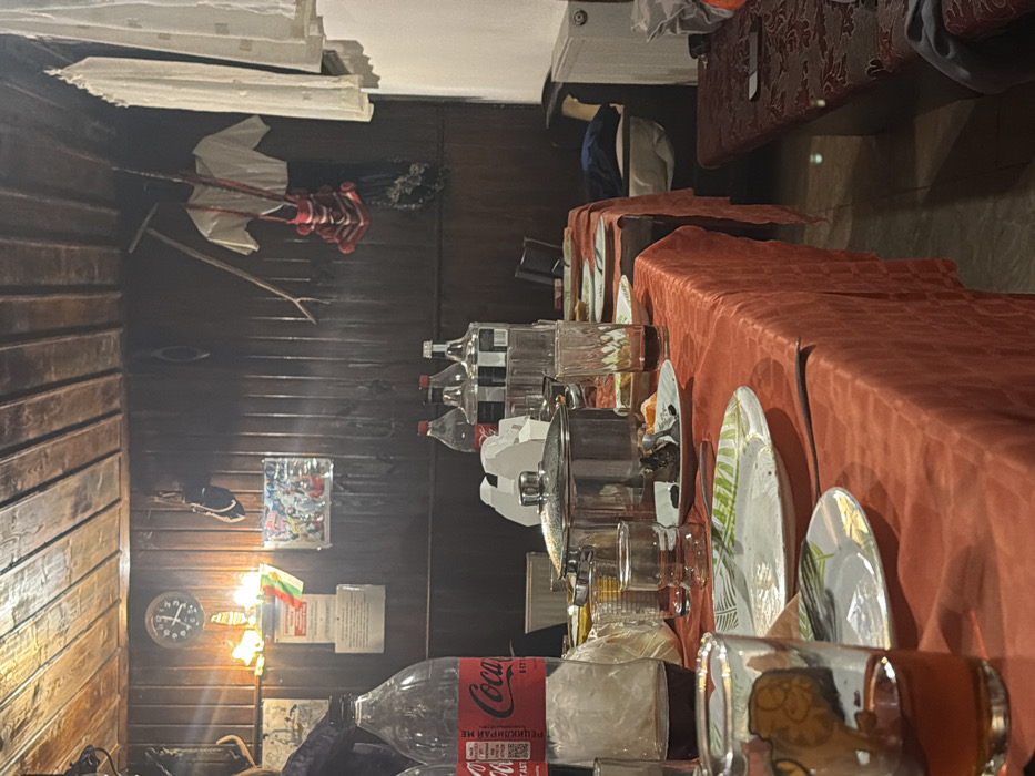 The image depicts an indoor scene in Turiya, Bulgaria, characterized by a rustic, traditional aesthetic, specifically a dining area after a meal. The room features dark wooden paneling on both the walls and the ceiling, with visible planks and joints.
A long dining table, covered with a textured orange-red tablecloth, occupies the foreground and midground. On the table, numerous items suggest a recent gathering: a large, silver lidded pot sits centrally, surrounded by several dirty white plates, some adorned with green and yellow leaf patterns. Food remnants, including dark pastry-like items and orange peels, are visible on some plates. Multiple clear drinking glasses, some containing residual red or amber liquid, are scattered across the table, along with cutlery. Two large plastic Coca-Cola bottles, one almost empty, stand on the left. To the right of the pot, at least three clear glass bottles with white labels contain a transparent liquid. A white cardboard carrier is also present.
The background wall is adorned with various objects. A round wall clock with a dark face and gold trim is positioned to the left. Below it, a bright wall-mounted light fixture with multiple bulbs illuminates the area, casting a warm glow. A banner displaying the Bulgarian flag colors (green, white, red) is affixed near the lamp, alongside a framed picture or poster featuring multiple colorful figures. Further right, a pair of dark shoes or slippers, a long wooden stick or tool, a light-colored traditional garment (possibly a blouse), a red woven bag with tassels, and a dark, possibly fan-like object are hung on the wall. To the far right, white curtains or fabric hang, and a white radiator is visible below.
No individuals are present in the frame, but the arrangement of dishes and beverages indicates a recent social event or meal. A dark wooden bench with a patterned cushion runs along the right wall, extending towards a partially visible armchair or couch further back.