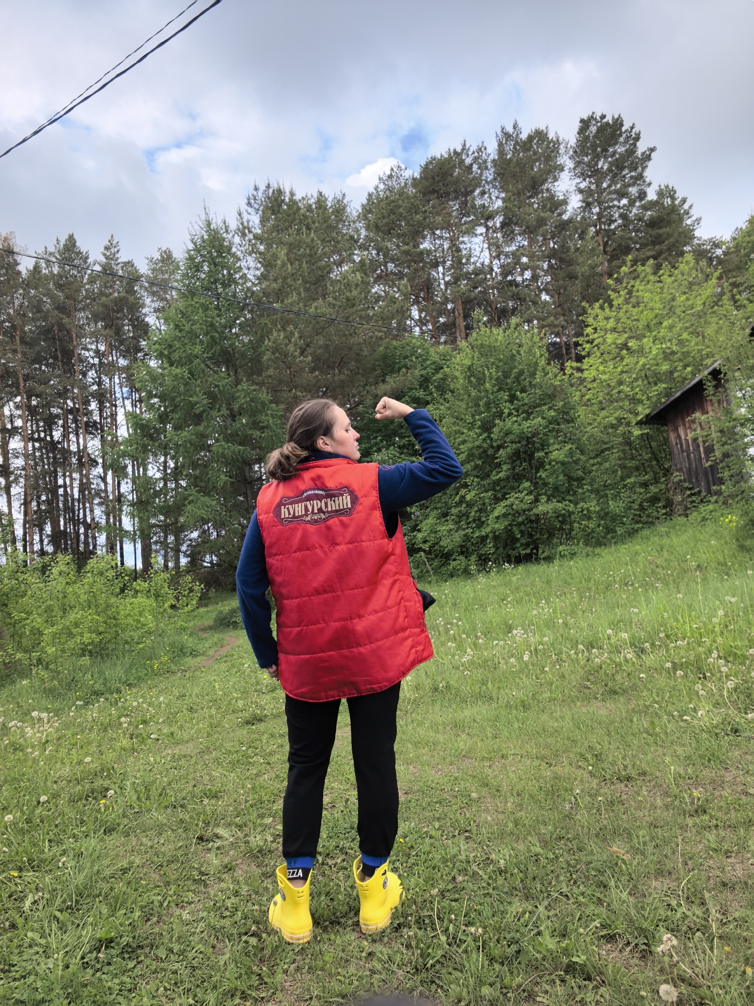 A person stands in a grassy field surrounded by trees, facing away from the viewer and flexing their left arm. They are wearing a red vest with Cyrillic text, black pants, blue socks, and bright yellow boots with "EZZA" printed on the side. The background features a mix of coniferous and deciduous trees, with a small wooden structure visible on a slight hill to the right. Above the trees, the sky is partly cloudy with visible power lines. The field is covered in green grass with white dandelions scattered throughout.