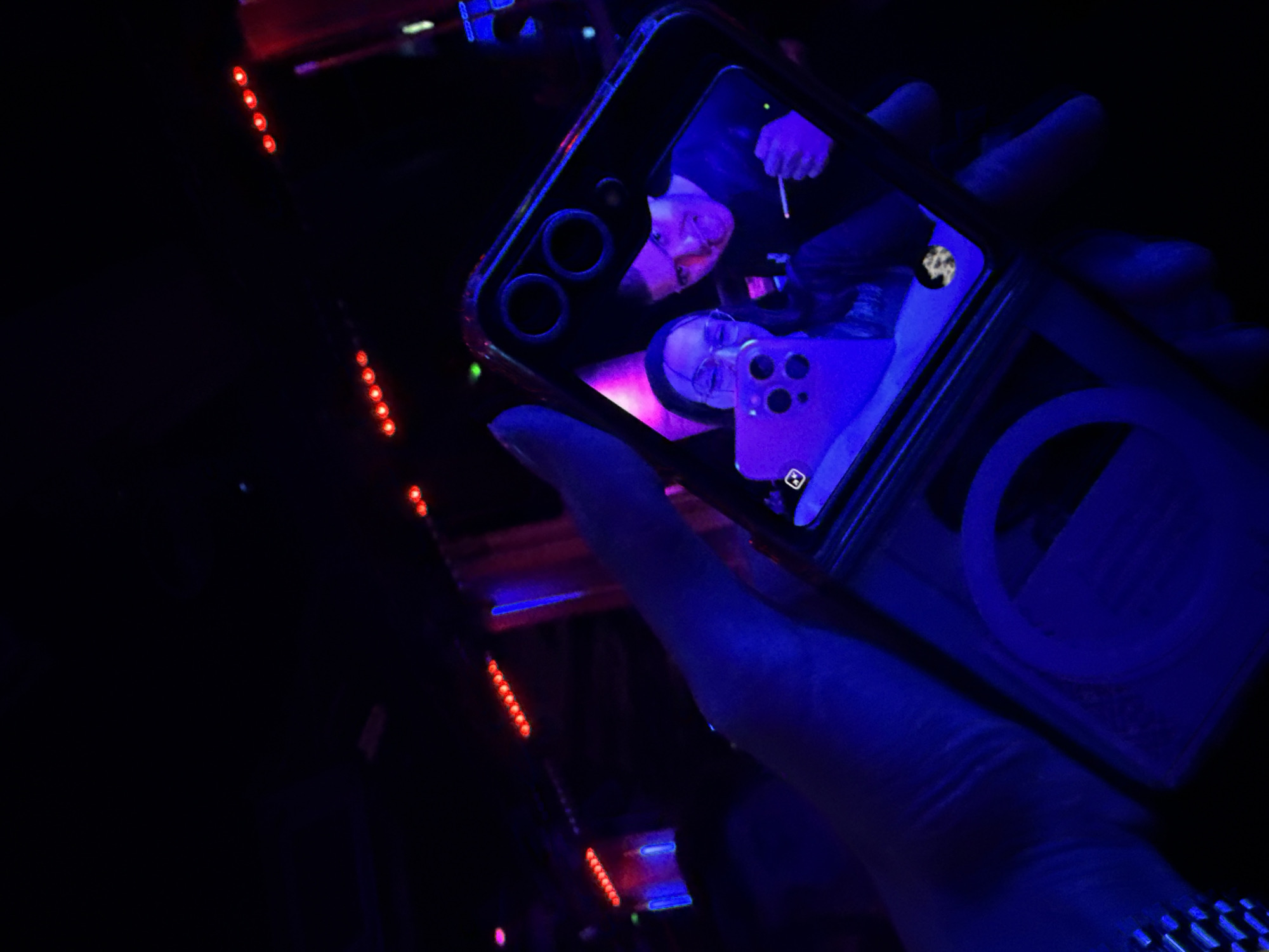 A person is holding a smartphone in their hand. The phone displays a screen recording of themselves, as well as another person with a cigarette. The background consists of red LED lights. The photo was likely taken in an entertainment venue, perhaps in Varna, Bulgaria, but this cannot be verified solely based on the image provided. The image is dominated by dark tones and bluish lighting.