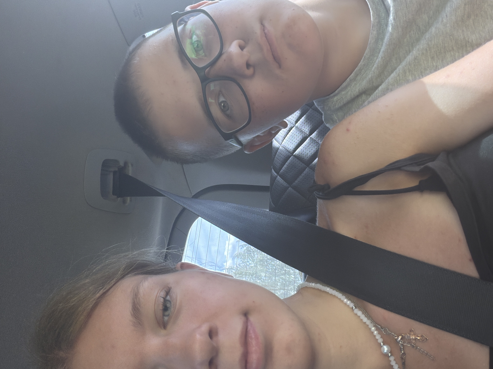 This is a selfie taken inside a vehicle, likely a car, with two young people visible. The person on the left, a female, is partially visible from the shoulders up. She has blonde hair and is wearing a black top and a beaded necklace with a cross pendant. The person on the right, a male, is wearing glasses and a grey t-shirt. He is also partially visible from the shoulders up. Both individuals appear to be looking towards the camera. A black seatbelt is prominently displayed across the foreground, dividing the two subjects. The background shows a quilted car seat and a window with trees and sky visible outside. The environment suggests a journey or outing. There is no specific indication of the location being Novoe Teneevo, Russia, within the image itself.