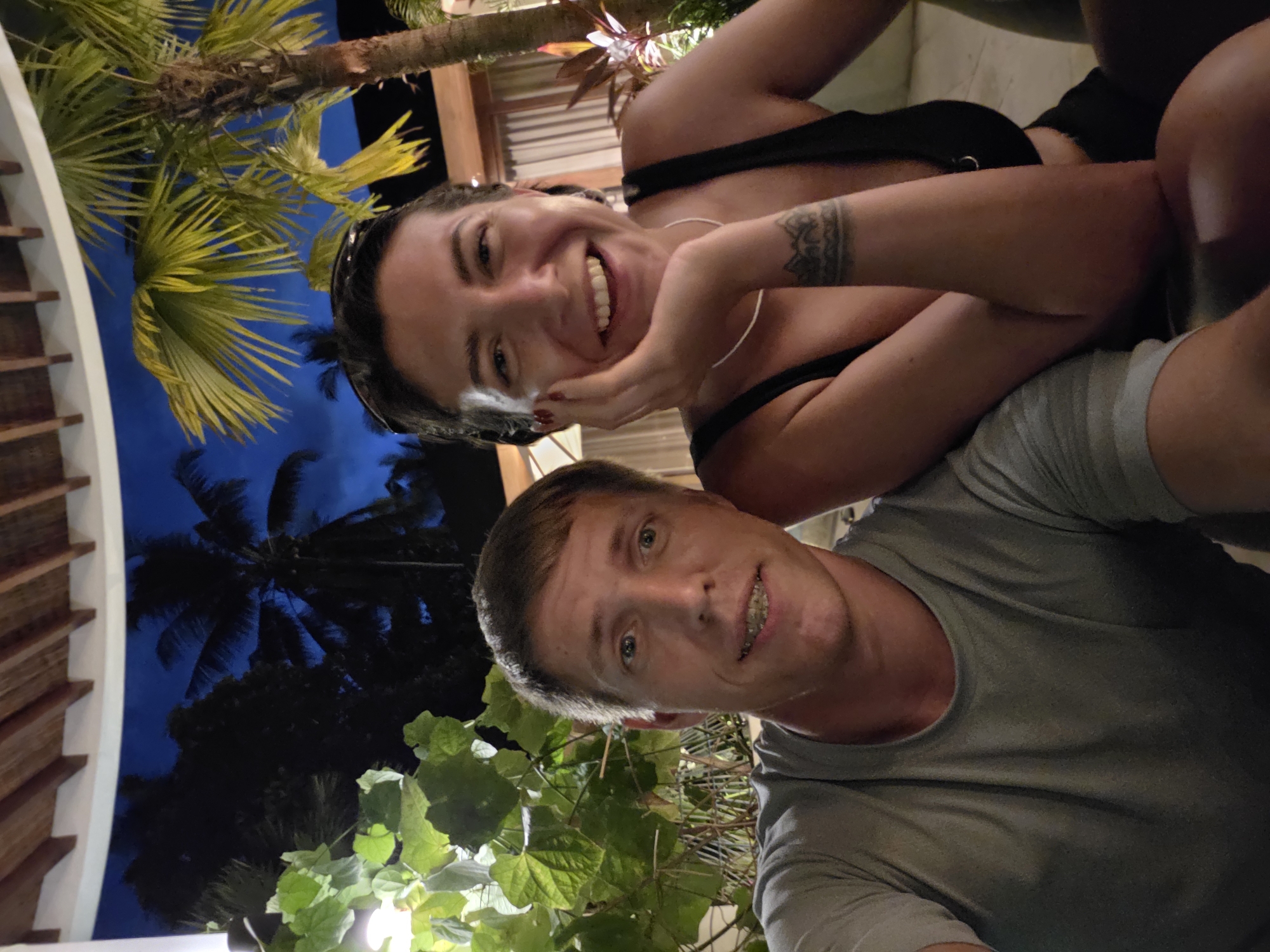 A low-angle shot shows a man and a woman posing for a selfie in what appears to be Ubud District, Indonesia. The woman, positioned behind the man, is smiling broadly, her hand resting on her cheek. She wears a black top and has a tattoo on her left forearm. The man, in the foreground, is wearing a grey t-shirt and also smiling, revealing braces.
Behind the couple, palm trees and lush green foliage are visible, lit by artificial lights, suggesting it is nighttime. The background includes a building structure with wooden beams, typical of the local architecture in Ubud. The dark blue night sky is visible between the palm trees.
