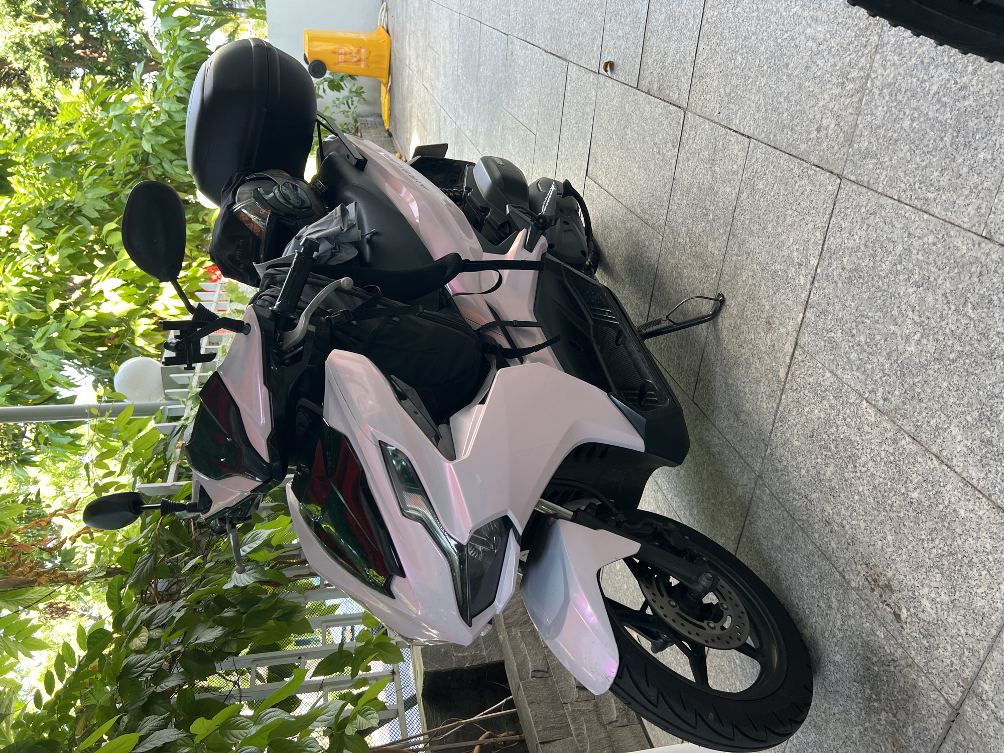 A white scooter is the central subject of this outdoor photograph, captured from a slightly elevated, front-side angle. The scooter features iridescent white body panels that subtly shift color, appearing light pink or purple in certain light, contrasting with its black seat, handlebars, and wheels. A black top case is mounted on the rear, and a black full-face helmet, along with several black bags and straps, are visible on or around the seat area, suggesting the vehicle is used for transport and carrying belongings. The front wheel, equipped with a disc brake, is partially visible.
The scooter is parked on a paved surface made of light grey, speckled granite-like tiles, which extend to a matching wall on the right side of the frame. To the left, abundant green foliage from trees and bushes creates a lush backdrop, suggesting a verdant environment. A white metal fence or railing is partially visible amidst the greenery. In the background, a bright yellow, rectangular object, possibly a utility box or container, is discernible.
The scene is brightly lit by natural sunlight, indicating it is daytime with clear weather. There are no people visible, and the scooter appears to be stationary, parked on its kickstand. The overall setting implies an outdoor, possibly urban or semi-urban location, consistent with the provided context of Nam Nha Trang Ward, Vietnam. No discernible text is visible in the image.