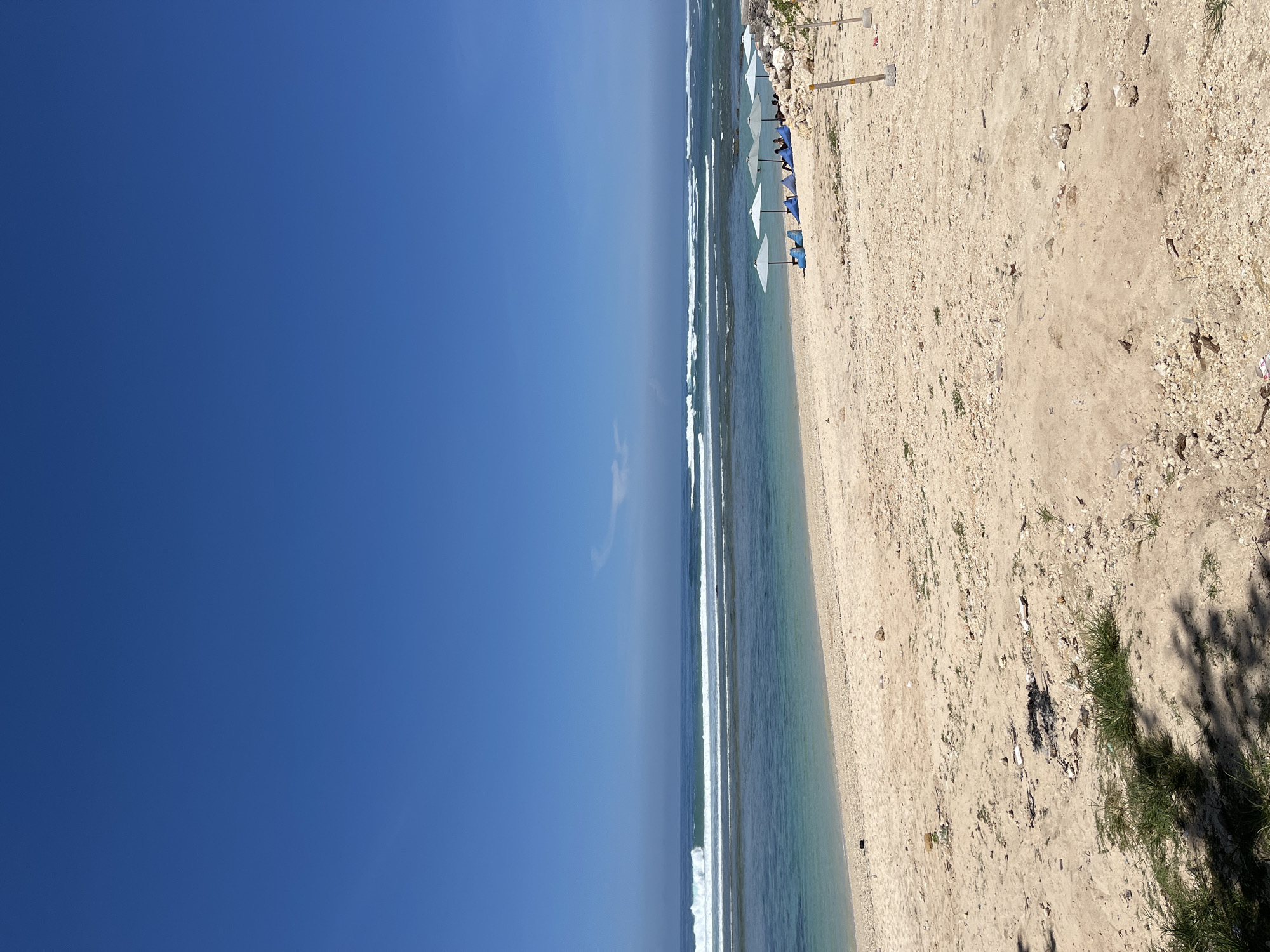 The image shows a beach in Kuta Selatan, Indonesia. The beach is sandy, with some small plants and debris scattered around. There are several beach umbrellas lined up along the shore. The ocean is calm with small waves. The sky is clear and blue.