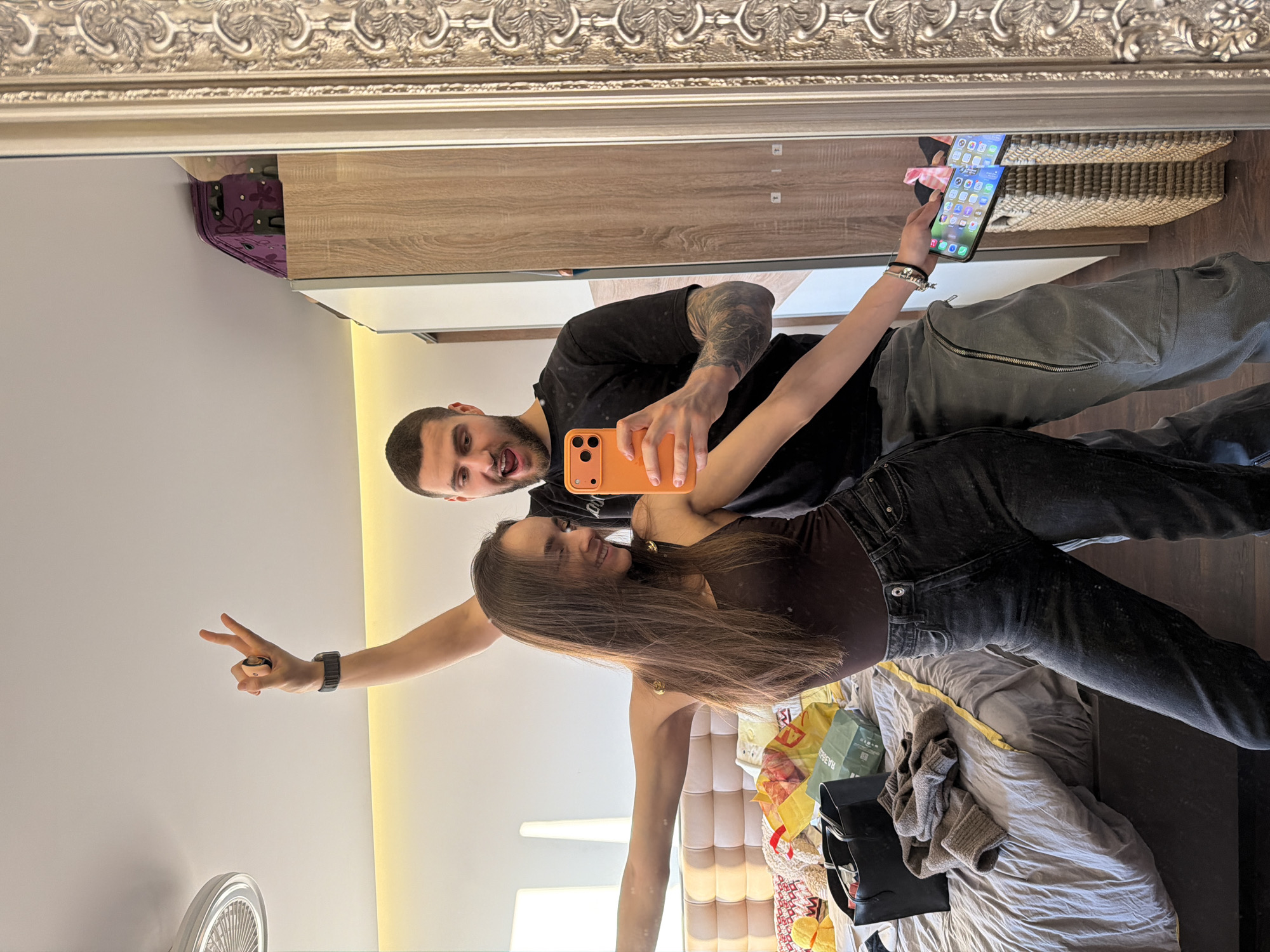 A couple takes a selfie in a mirror. The man, fair-skinned with a beard, dark hair, and tattoos on his left arm, holds an orange phone to capture the image. He wears a black t-shirt and grey pants. The woman, with long brown hair, a brown top, and dark jeans, stands beside him. She holds up two fingers in a peace sign with her left hand. The reflection in the mirror shows a room with a bed covered with a grey comforter, shopping bags, and personal belongings. There's also a fan in the corner and a light fixture above the bed. Behind them is a wardrobe. The city and specific location in Sofia, Bulgaria, are not evident from the image.