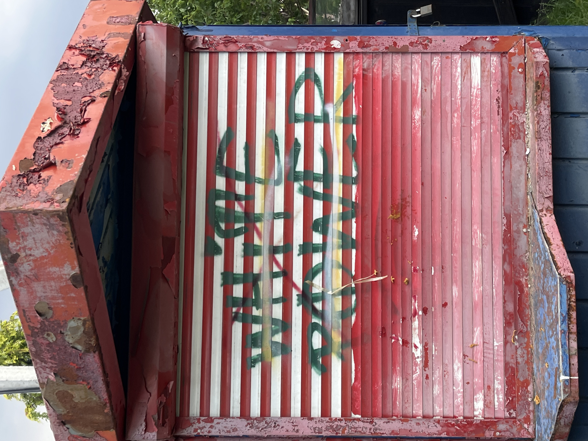 The image shows a weathered red kiosk with a metal roll-up door. Graffiti in green and yellow spray paint is visible on the door, reading "WHITE POWER". The kiosk appears to be in a state of disrepair, with peeling paint and rust. The surrounding environment suggests an outdoor urban setting, with some greenery visible in the background. The kiosk is located on a street in Minsk, Belarus.