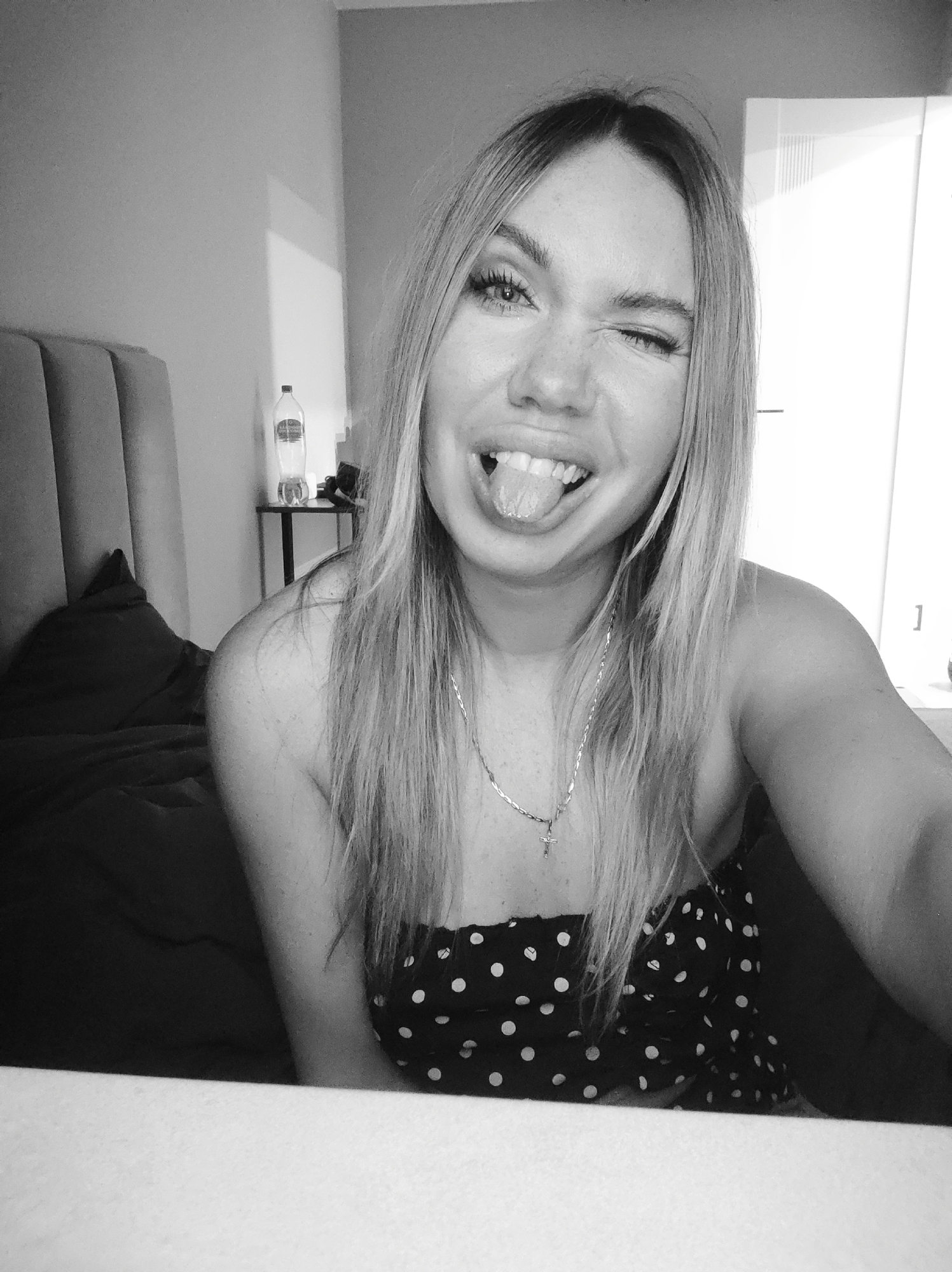 A black and white selfie shows a woman with long blonde hair, wearing a polka-dot top and a necklace with a cross pendant, winking and sticking out her tongue. She is indoors, possibly in a bedroom, as evidenced by a bed with a headboard behind her. A small table with a water bottle on it is visible in the background. There is no evidence of the city of Votkinsk, Russia.