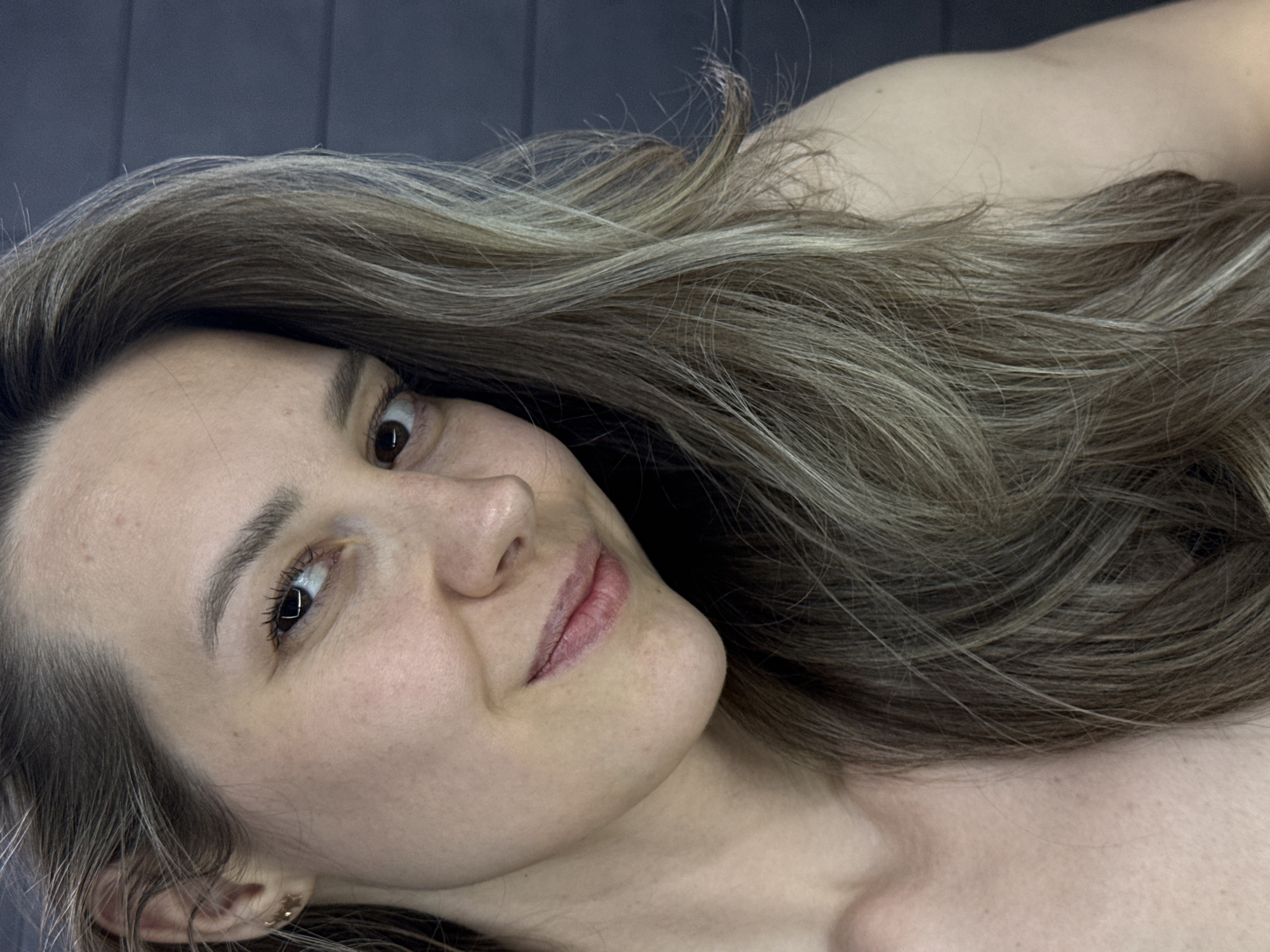 A headshot of a woman with light brown hair streaked with blonde highlights is centered in the frame. Her face is turned towards the camera, with a slight smile. Her eyes are brown, and she appears to be wearing mascara. The background is a grey, textured surface. A small gold earring is visible in her ear. Her bare shoulder and chest are visible in the lower right corner. The location is not discernible from the provided image. The woman's name, any action or interaction, or the environment is not discernible. The image is oriented horizontally.