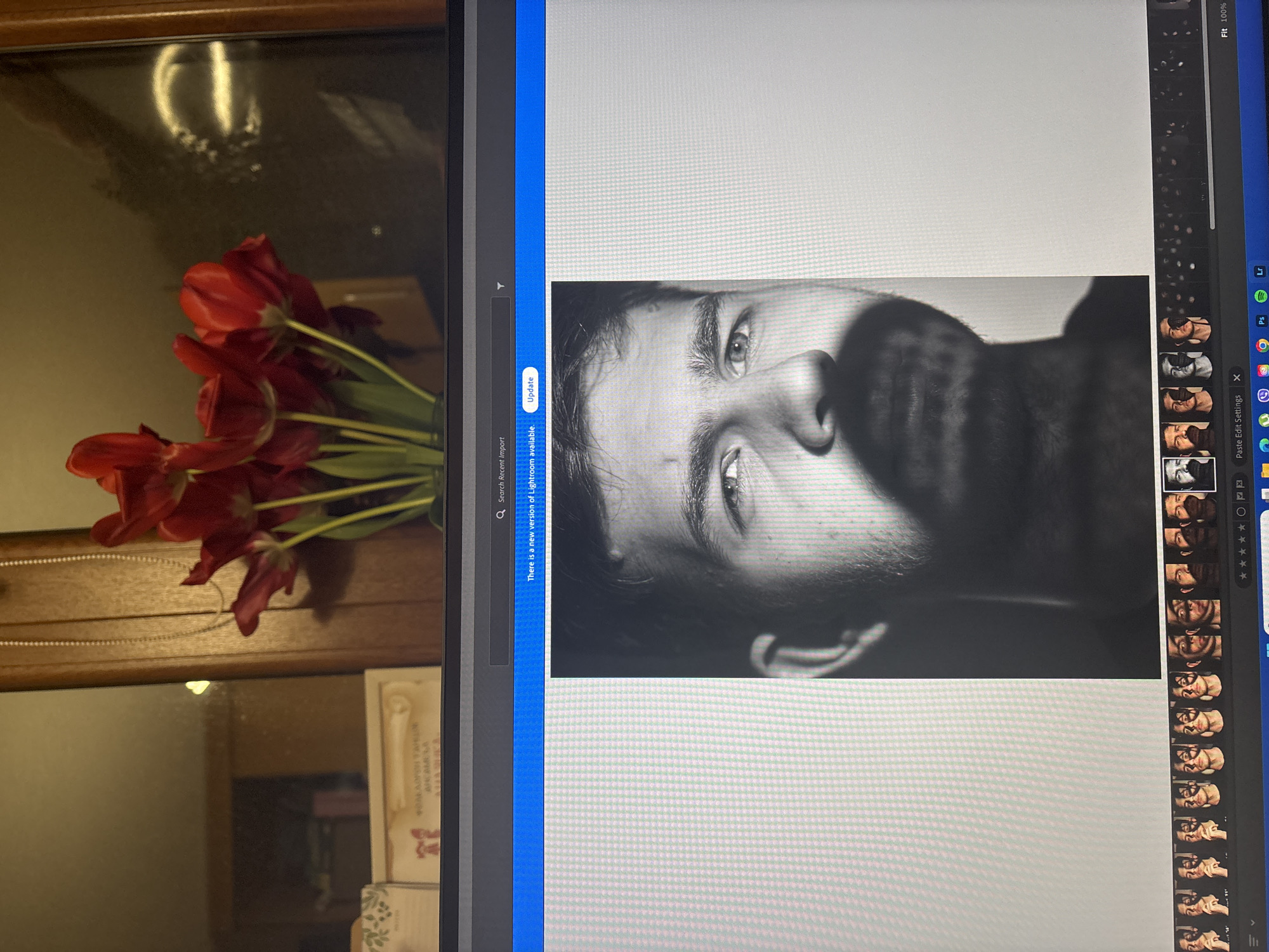 The image shows a juxtaposition of two scenes. On the left, a bouquet of red tulips rests on a wooden surface in front of a mirror. The mirror reflects a blurred interior scene. On the right, a computer screen displays a close-up, black and white portrait of a man with light eyes and stubble. The screen also shows the Lightroom interface with a panel of image thumbnails.
There is nothing in the image to suggest the location is in Plovdiv, Bulgaria.
