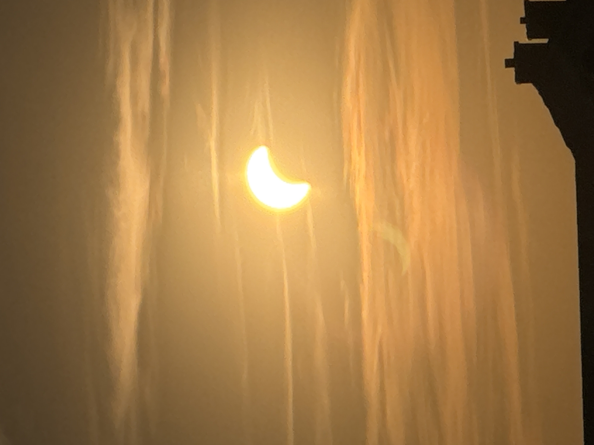 A partial solar eclipse is visible in a hazy, orange sky. The sun, partially obscured, appears as a crescent shape. Streaky clouds are visible throughout the sky, and the silhouette of rooftops with chimneys is visible at the bottom of the image. This image was taken in Wettstetten, Germany.