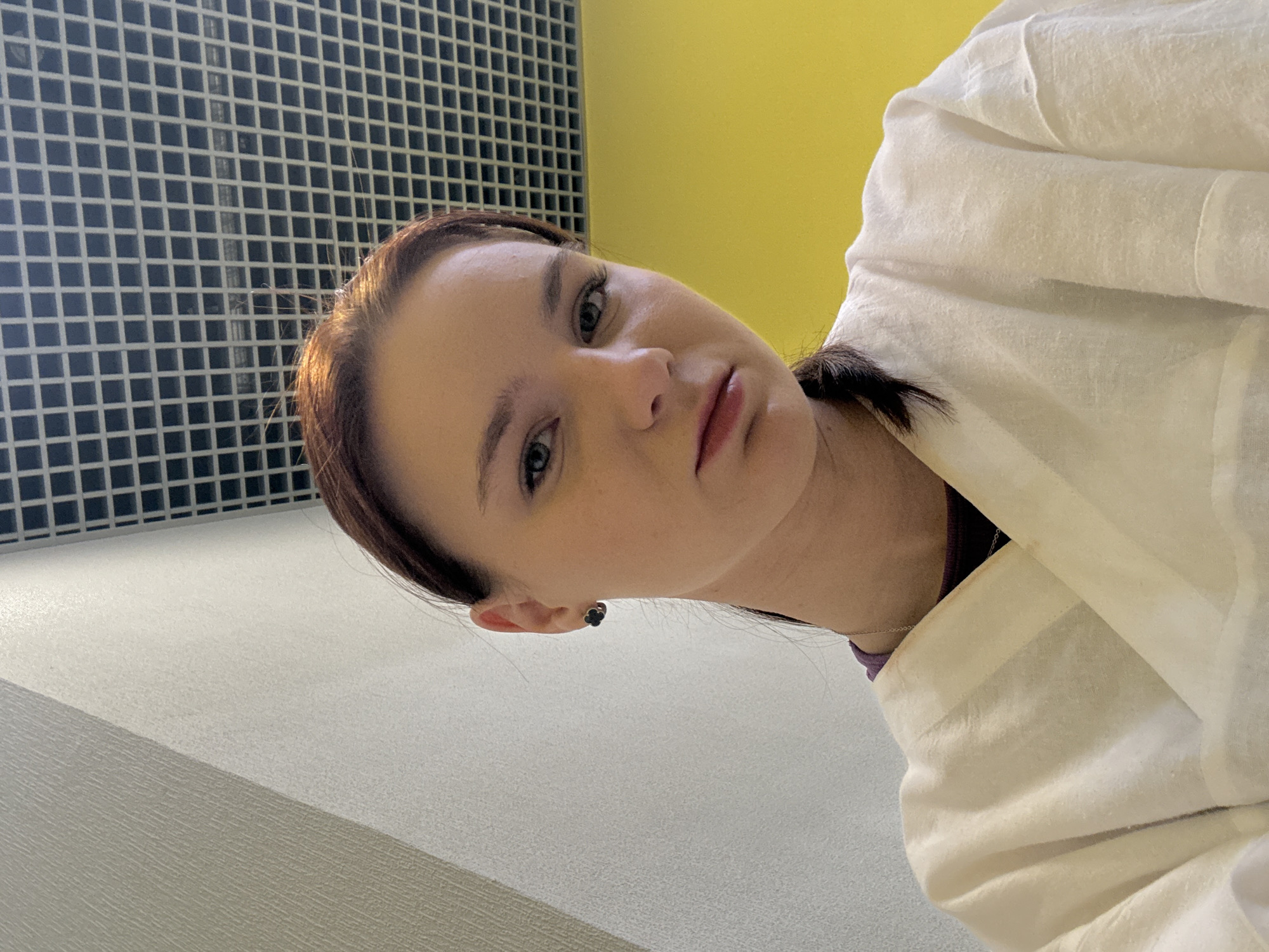 A woman with light skin, dark hair pulled back, and wearing a white garment stands indoors. She is angled towards the camera with a slight tilt of her head. Behind her is a grid-like structure of dark squares on a light background and a plain yellow wall. It's not possible to confirm the location in Sinyavino, Russia, based on the image alone.