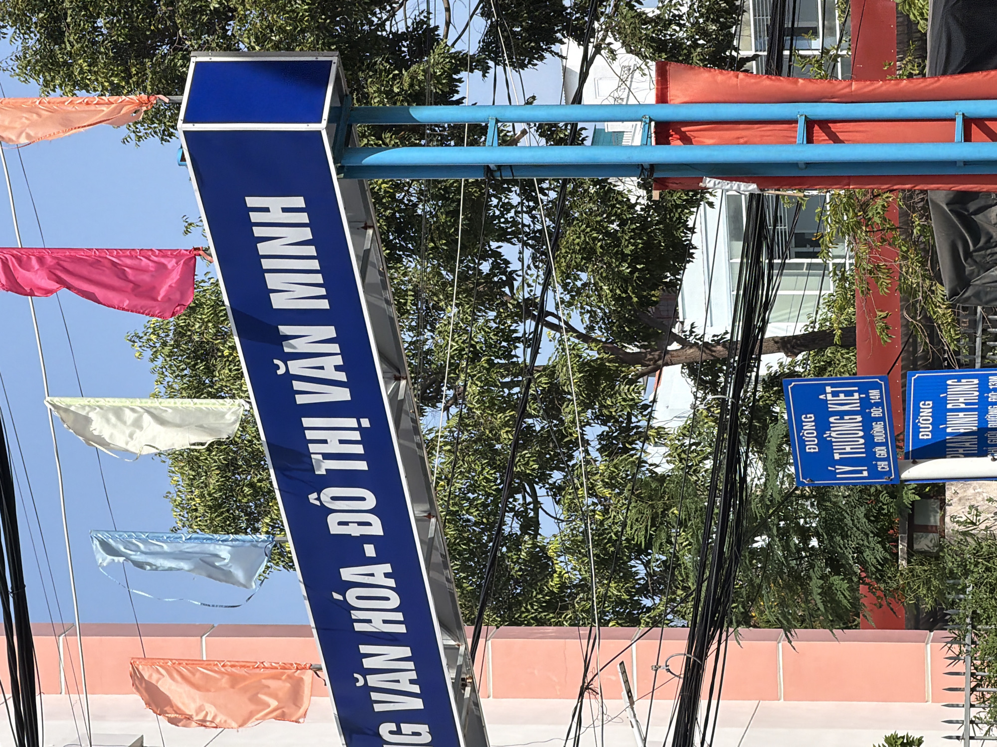 The image shows a street scene in Nha Trang, Vietnam, focusing on a blue sign with white lettering and several colorful flags. The sign reads "TRUNG TÂM VĂN HÓA - ĐÔ THỊ VĂN MINH". Below this, two blue street signs are visible. The upper sign indicates "ĐƯỜNG LÝ THƯỜNG KIỆT" and "CHỈ GIỚI ĐƯỜNG: 14M". The lower sign reads "ĐƯỜNG PHAN ĐÌNH PHÙNG" and "CHỈ GIỚI ĐƯỜNG: 13M".
Several flags are attached to poles above the main sign. From left to right, there is an orange flag, a light blue flag, a white flag, and a pink flag, followed by another orange flag on the right. A tall structure with two blue poles supports the main sign, and a red banner is attached to it. The background features green trees, a light pink building on the left, and a white building with large windows in the distance. Numerous black electrical wires are strung across the scene. The sky is clear and blue. No people are visible in the image.