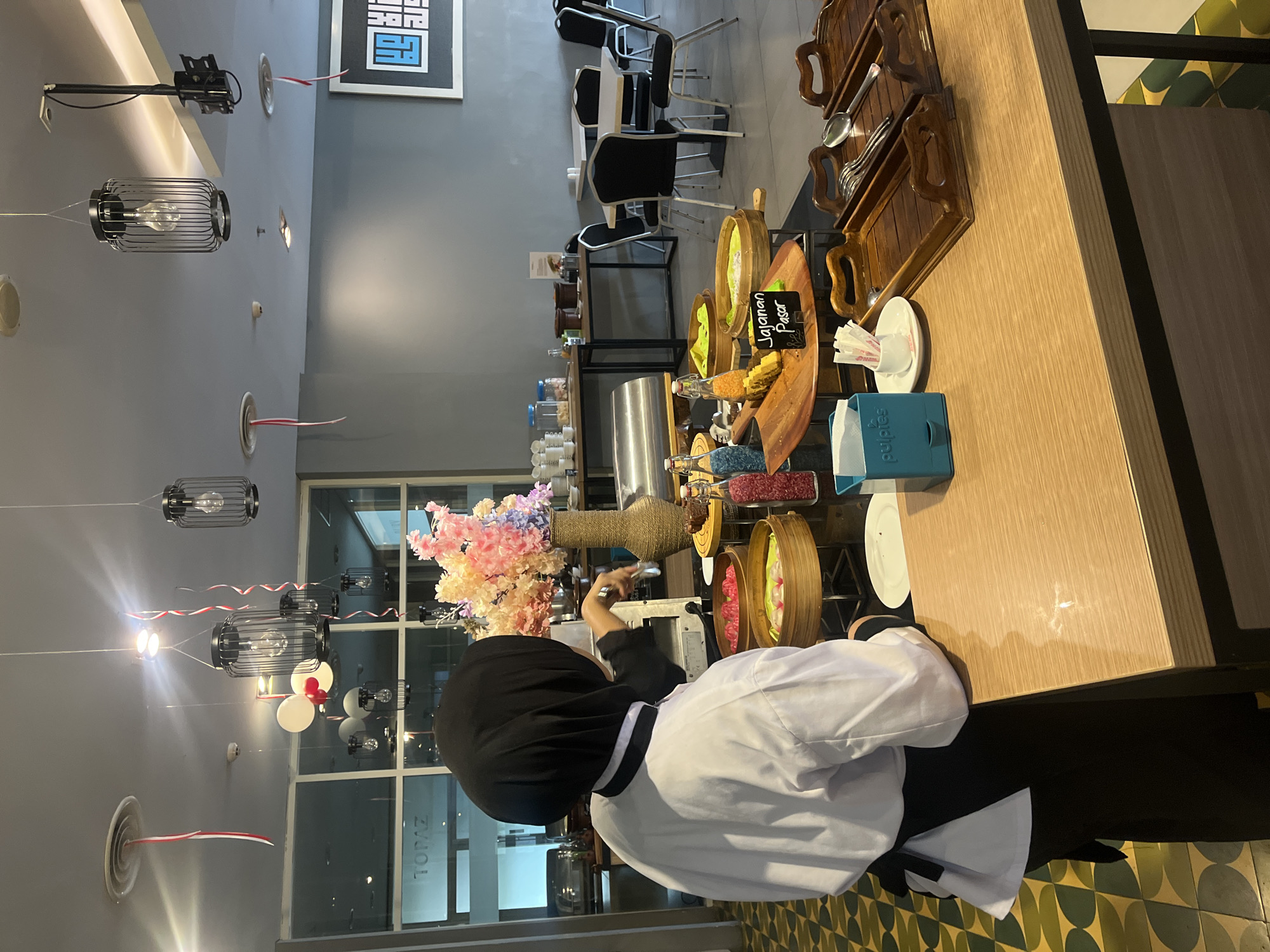 The image depicts a buffet setting inside a modern restaurant, possibly a hotel, in Gapurosukolilo, Indonesia. A person wearing a black hijab and a white chef's uniform with a black apron is seen from the back, serving food. They are standing behind a wooden counter displaying various food items in bamboo steamers and glass bottles containing colorful ingredients. To the right of the food display, there are wooden trays holding cutlery and a blue tissue dispenser. In the background, tables and chairs are visible, along with large windows that offer a glimpse of the outdoors or another section of the building. Decorative elements like lanterns hanging from the ceiling and streamers add to the ambiance. The overall environment suggests a meal service, possibly breakfast or brunch, with an emphasis on a diverse culinary offering.