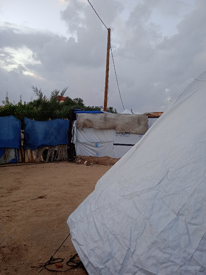 A low-angle shot captures an outdoor scene with a large white tent dominating the foreground, its folds and wrinkles visible. Behind it, makeshift shelters stand, constructed from materials like blue fabric, straw, and white plastic. A tall wooden pole rises beside these structures, extending towards a cloudy sky. The ground is sandy and uneven. The composition suggests a temporary or impoverished setting.