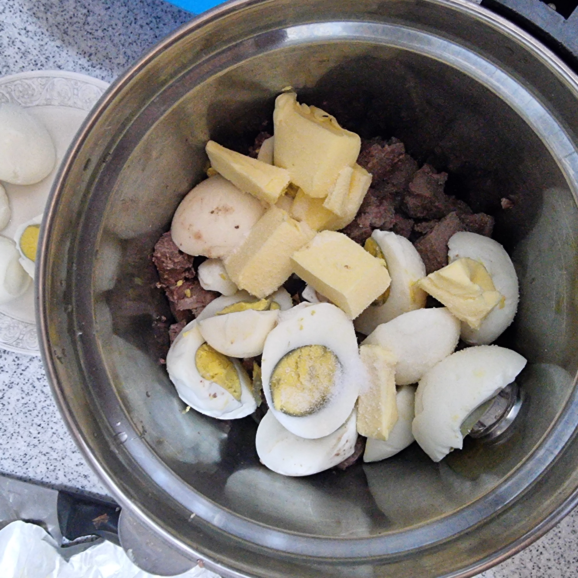 The image shows a close-up, overhead view of a stainless steel bowl containing chopped hard-boiled eggs and what appears to be ground meat. The eggs are mostly halved or quartered, revealing yellow yolks. Some of the egg pieces have a dusting of white, granular material, likely salt. Interspersed with the eggs are chunks of a pale yellow substance, possibly butter or cheese. The ground meat is a dark reddish-brown. The bowl is placed on a speckled gray surface, which might be a countertop. To the left of the bowl, a crumpled white plastic bag and a portion of a black object are visible. In the background, a white plate with several more hard-boiled eggs, some whole and some halved, is visible on the same speckled surface. The overall impression is of ingredients being prepared for a meal. The image is undated and no specific location information is available.