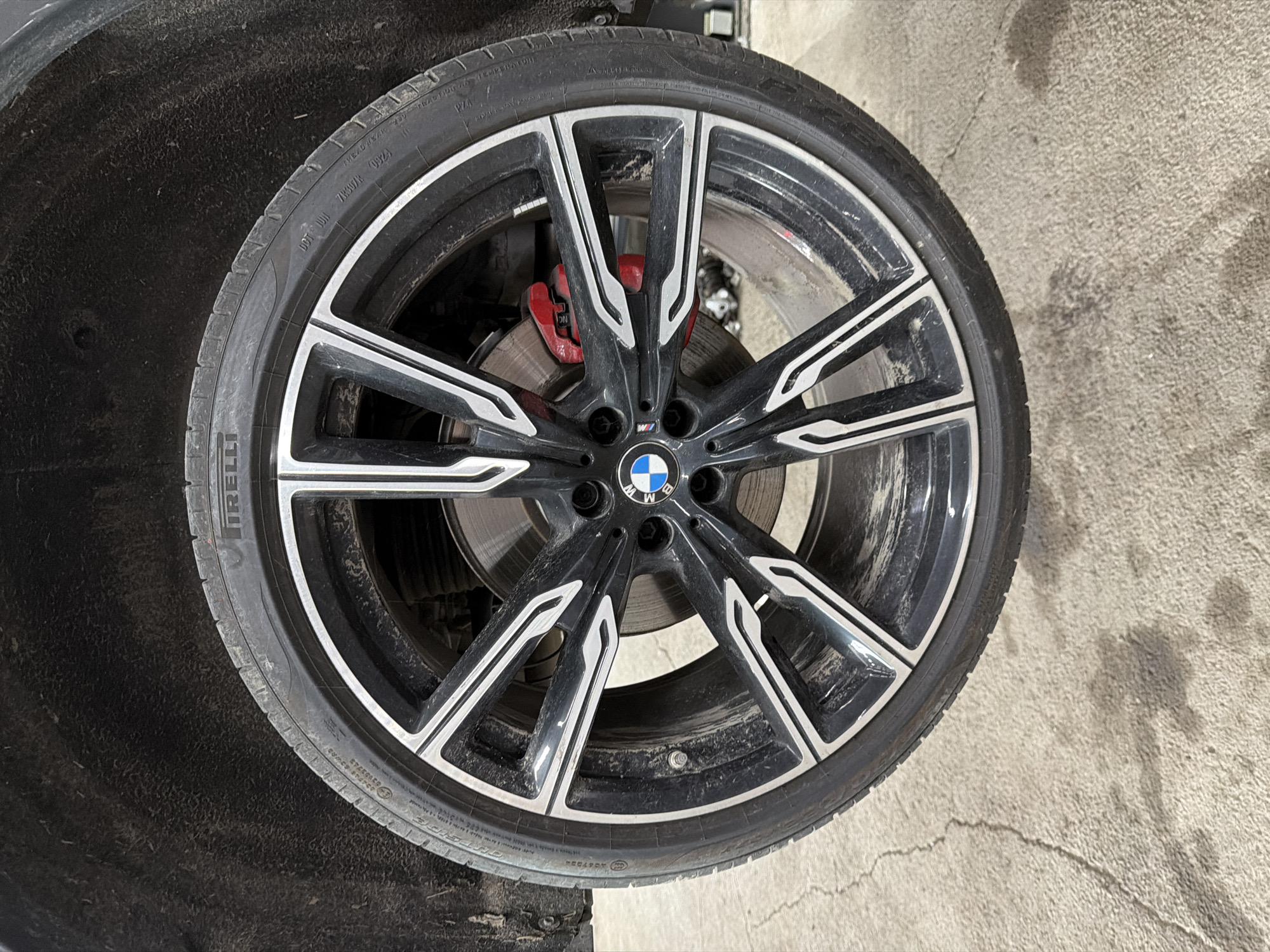 The image presents a close-up view of a single automotive wheel, likely from a BMW, set within a garage or workshop context in Burgas, Bulgaria. The wheel consists of a black multi-spoke alloy rim with contrasting polished or silver accents on the spokes. A circular BMW logo, featuring blue and white quadrants, is centered on the hub, with an "M" logo also visible on one of the spokes.
A Pirelli P Zero tire is mounted on the rim. Visible text on the tire sidewall includes "PIRELLI," "P ZERO," "TREADWEAR 200," "TRACTION A," "TEMPERATURE A," "DOT AR307R 092A," and the size specification "225/40R19 93Y." Additional markings like "M+S" and "EXTRALOAD" are also discernible. The tire and rim show signs of use, with accumulated dirt and dust.
Through the spokes of the rim, a bright red brake caliper is prominent, gripping a metallic brake disc. The caliper features a stylized "M" logo, indicating it's part of a BMW M performance braking system. The brake disc exhibits typical wear patterns.
The immediate background to the upper left is a dark, textured surface, consistent with a vehicle's inner fender well or wheel arch. The foreground and right side of the image reveal a light gray concrete floor, characterized by cracks and stains, typical of a service area. The scene suggests the wheel is either being prepared for installation, removed from a vehicle, or undergoing inspection.