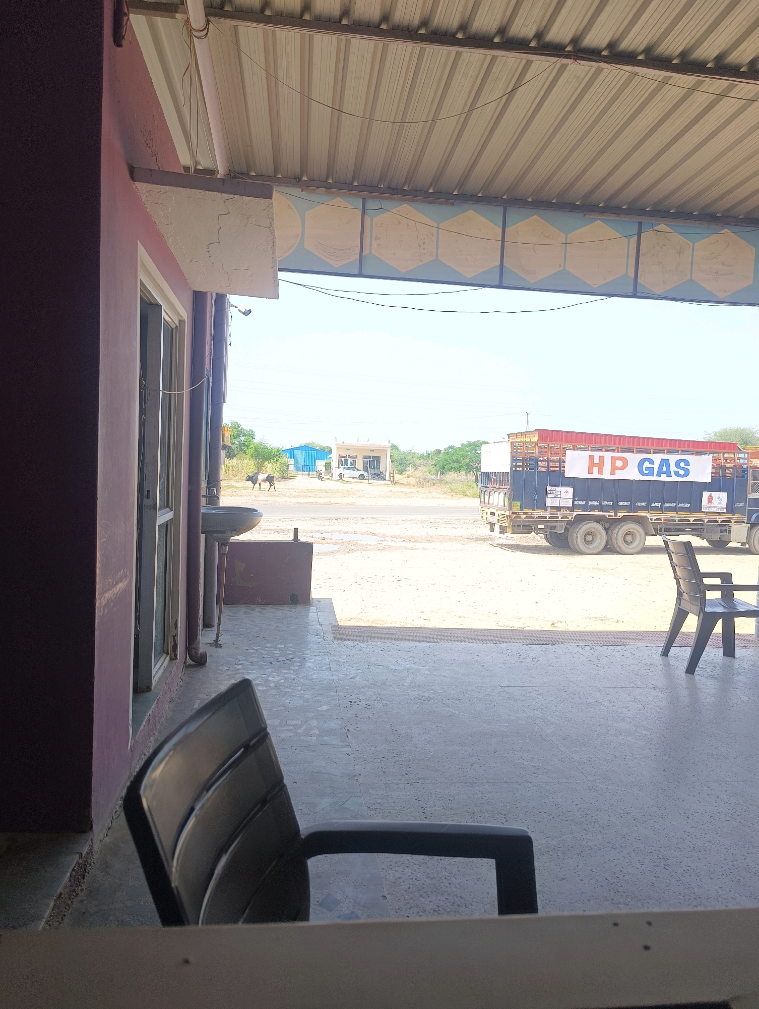 The image depicts an outdoor scene in Chhapri Bari, India, viewed from under a covered patio. In the foreground, a dark plastic chair is visible, positioned on a grey, speckled tiled floor. To the left, the corner of a building with a maroon-colored wall and a white window frame is present. Beyond the patio, a dusty road is visible, with a large blue and red truck parked on it. The truck has "HP GAS" printed in white letters on an orange banner on its side. In the distance, a blue building and a white car are visible, along with some dry vegetation and utility poles under a clear, pale blue sky. A lone bovine animal stands near the road in the mid-ground.