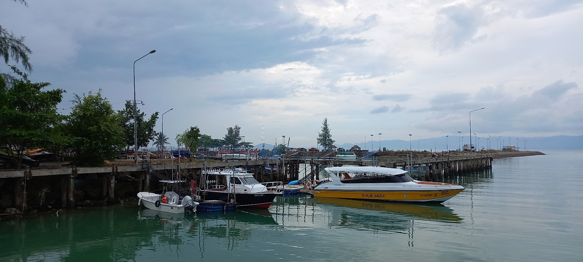 The image depicts a harbor in Baan Tai, Thailand, under an overcast sky. Three boats are docked along a weathered concrete pier lined with evenly spaced street lamps. From left to right, there is a small white boat, a larger black and white boat, and a yellow speedboat bearing the name "KHUN ANDA." The boats are reflected in the calm, greenish water. The pier extends into the water, with various vessels docked beyond. Trees line the shoreline behind the pier. The background shows a distant coastline under the cloudy sky.