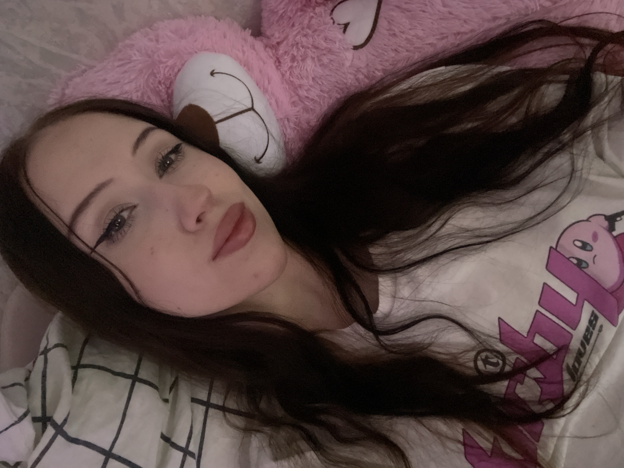 A young woman with long, dark hair lies in bed next to a large pink teddy bear. She is wearing a white t-shirt with a pink graphic design on it. Her eyes are open, and she is looking at the camera. The room appears to be dimly lit. No specific location in Votkinsk, Russia, can be determined from the image.
