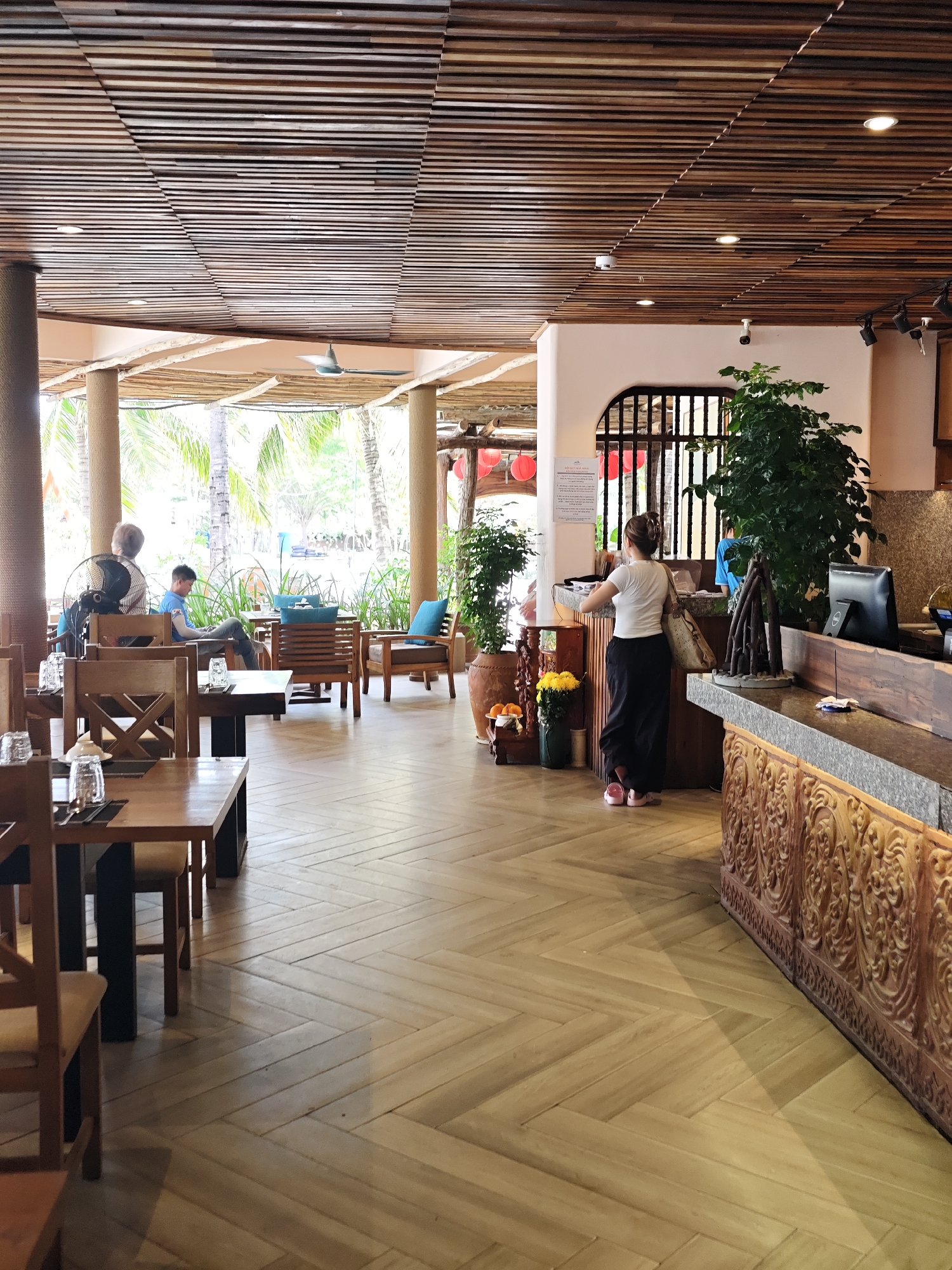 The image depicts the interior of a restaurant or cafe with an outdoor seating area visible through large windows. The architecture features a slatted wooden ceiling and a herringbone patterned wooden floor. In the foreground, several wooden tables and chairs are set, some with glassware and place settings. To the left, a large fan is positioned behind two patrons seated at a table. Further back, another patron is seated at a table with blue cushions. On the right side of the image, a woman in a white top and black pants is standing at a counter, interacting with a cashier or service staff, who is partially visible behind the counter. The counter itself is made of stone with ornate wooden carvings at its base. Plants are placed strategically throughout the space, including a large potted plant near the counter. Red lanterns are visible hanging in the background, suggesting a thematic or decorative element. The overall ambiance is relaxed and tropical, likely reflecting the coastal location of Phan Rang – Tháp Chàm, Vietnam, which is known for its beaches and resorts. The natural light suggests it is daytime.