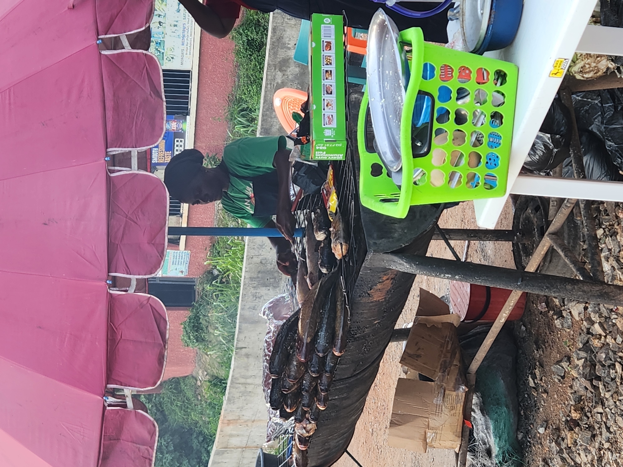 The image depicts an outdoor food stall in Jos, Nigeria, under a large pink umbrella. A woman wearing a green t-shirt and a dark head covering is tending to a grill laden with numerous fish. The fish are arranged on metal racks over a large, dark, oval-shaped charcoal grill. The scene suggests a busy street food operation. In the foreground, a bright green plastic basket holds metal plates and utensils, placed on a white table. Behind the grill, a reddish-brown building is visible with some signage. The ground is covered in gravel and dirt, with various items like cardboard boxes, a red bucket, and a large plastic bag scattered around. The overall atmosphere is that of a typical street vendor setup in a bustling urban environment.