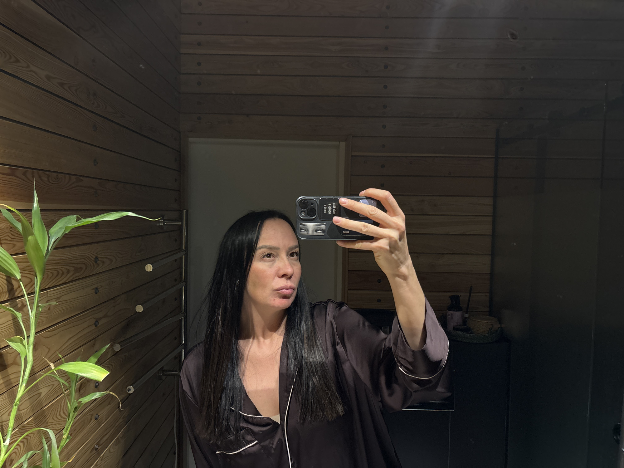 A person is taking a selfie in what appears to be a bathroom. The bathroom has a wooden plank wall and a bamboo plant in the lower left corner. The person is holding a smartphone with a black case up to take the picture. The person has long, dark hair and is wearing a dark-colored robe with white piping. A white door is visible behind the person. In the background to the right, there is a black cabinet or shelf. There is also what appears to be a towel rack attached to the wall. It is not possible to determine the exact location in the city of Chaikovsky, Russia, based on this image.