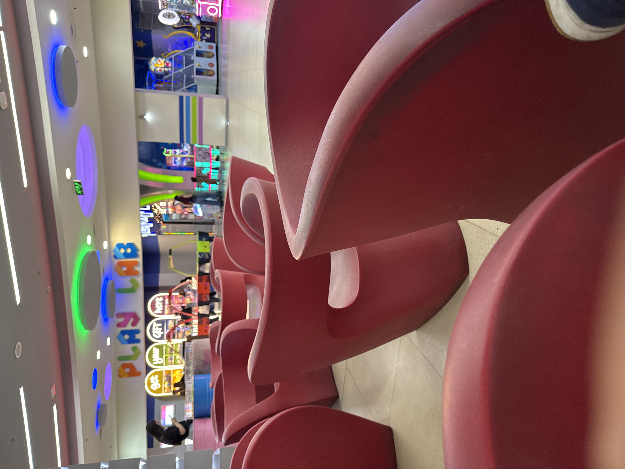 The image, oriented vertically, captures a vibrant indoor entertainment center, possibly within a mall in Yerevan, Armenia. The foreground and midground on the right and top are dominated by several rows of uniquely shaped, deep red chairs with a modern, sculptural design. In the extreme top right corner, a person's foot, wearing a blue and white sneaker, rests on one of these chairs.
The lower and left portions of the image reveal the brightly lit arcade area. A large, colorful sign for "PLAYLAND" is prominently displayed on a wall, oriented vertically with its letters stacked from bottom to top, illuminated in yellow, orange, green, and blue. Below this, three vertically stacked circular signs read "get," "your," and "GIFT here." The ceiling features various circular light fixtures glowing in blue, purple, and green, alongside white strip lighting.
Numerous arcade machines with flashing lights and screens are visible, creating a dynamic atmosphere. One person is seen in the bottom-left corner, seated and looking at their phone. The scene is well-lit by artificial lights, making the time of day ambiguous. The overall impression is one of a contemporary and inviting leisure space.