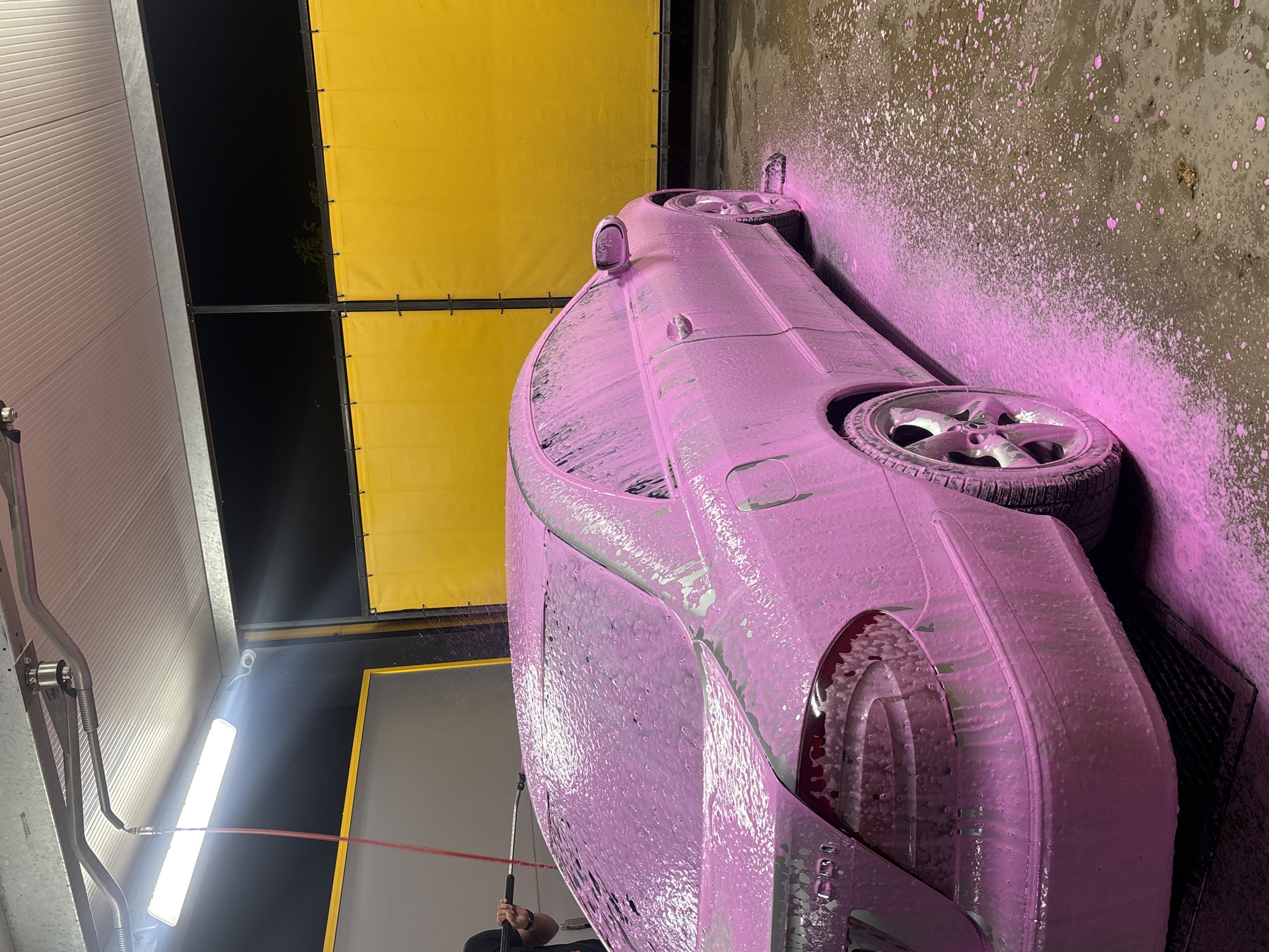 A silver sedan, covered in bright pink foam, is being washed at an outdoor self-service car wash. The car is positioned on a dark gray mat on the right side of the frame, with its rear towards the viewer. The foam covers the entirety of the vehicle, including the windows, wheels, and mirrors. The background features bright yellow curtains or dividers, and a dark night sky is visible above. A person's arm and hand, holding a black pressure washer wand, are visible on the left side of the image, actively spraying the car. The ground is wet and covered with scattered pink foam. The scene is illuminated by overhead lights. This car wash is located in Pleven, Bulgaria.