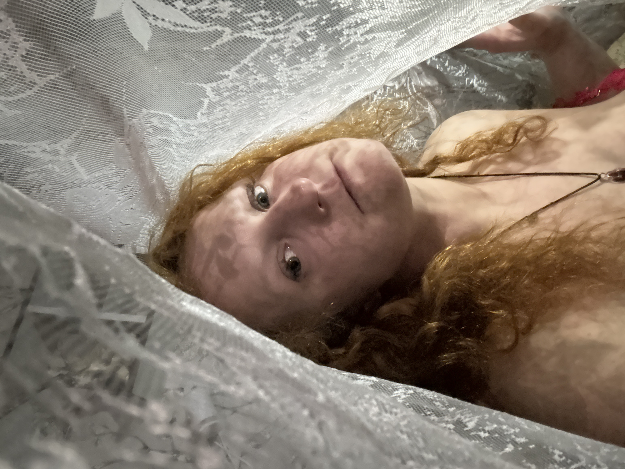 A woman with red hair lies on her back, partially covered by a white lace fabric. She wears a necklace. The setting's specific location in the city of Chaikovsky, Russia, cannot be determined from the visual data provided.