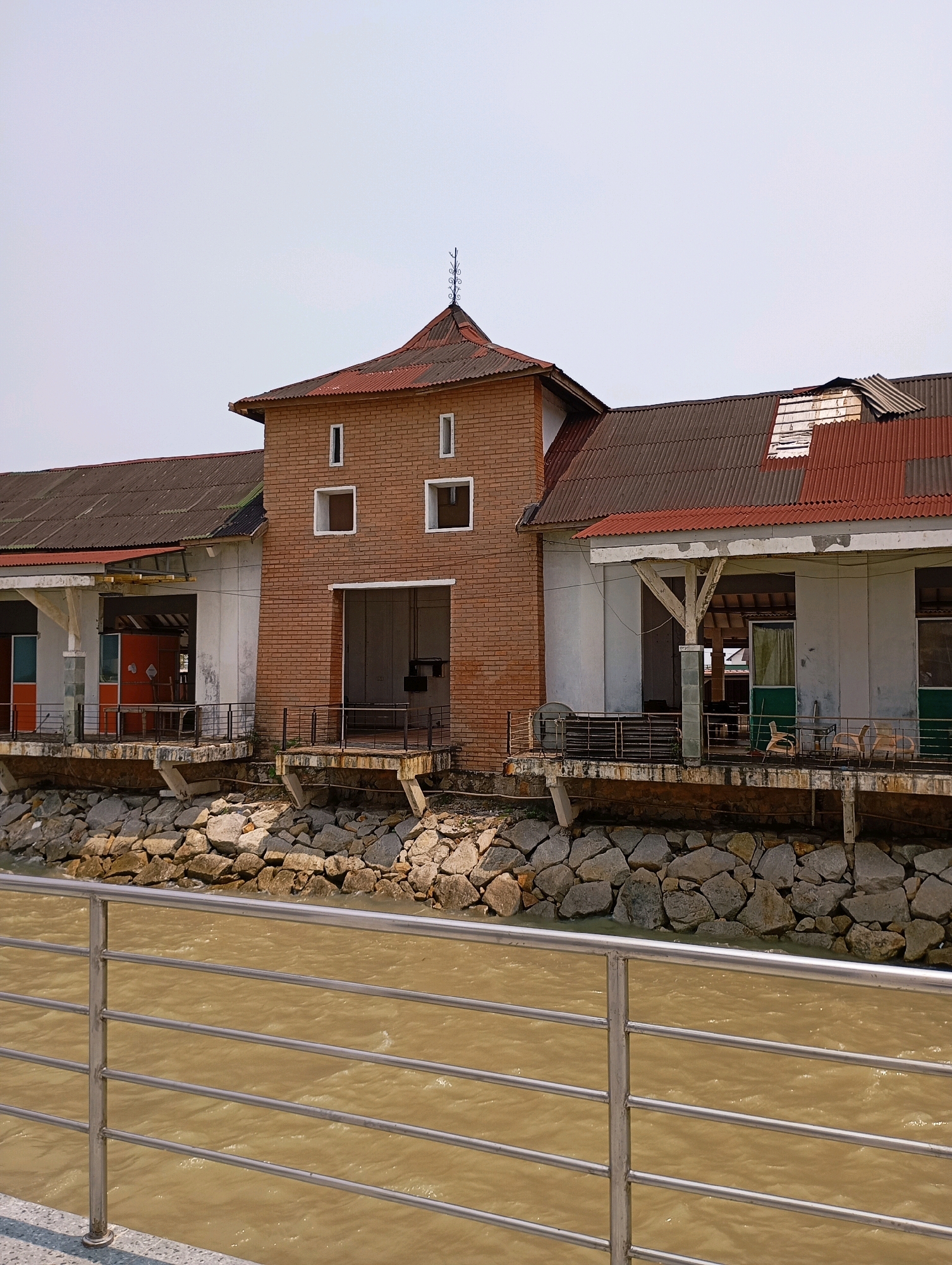 The image depicts a waterfront building with a complex roof structure and brick facade, situated in the city of Tanjung Balai Karimun, Indonesia. The building exhibits signs of age and disrepair, with corrugated metal roofing showing damage in places. It overlooks a murky, brownish body of water, likely a river or sea, protected by a rocky embankment. In the foreground, a modern metal railing runs horizontally across the bottom of the frame, suggesting a walkway or viewing area. The sky is a clear, pale blue. No people are visible in the scene. The architectural style suggests a utilitarian or possibly historical structure, possibly a former warehouse or dockside facility.