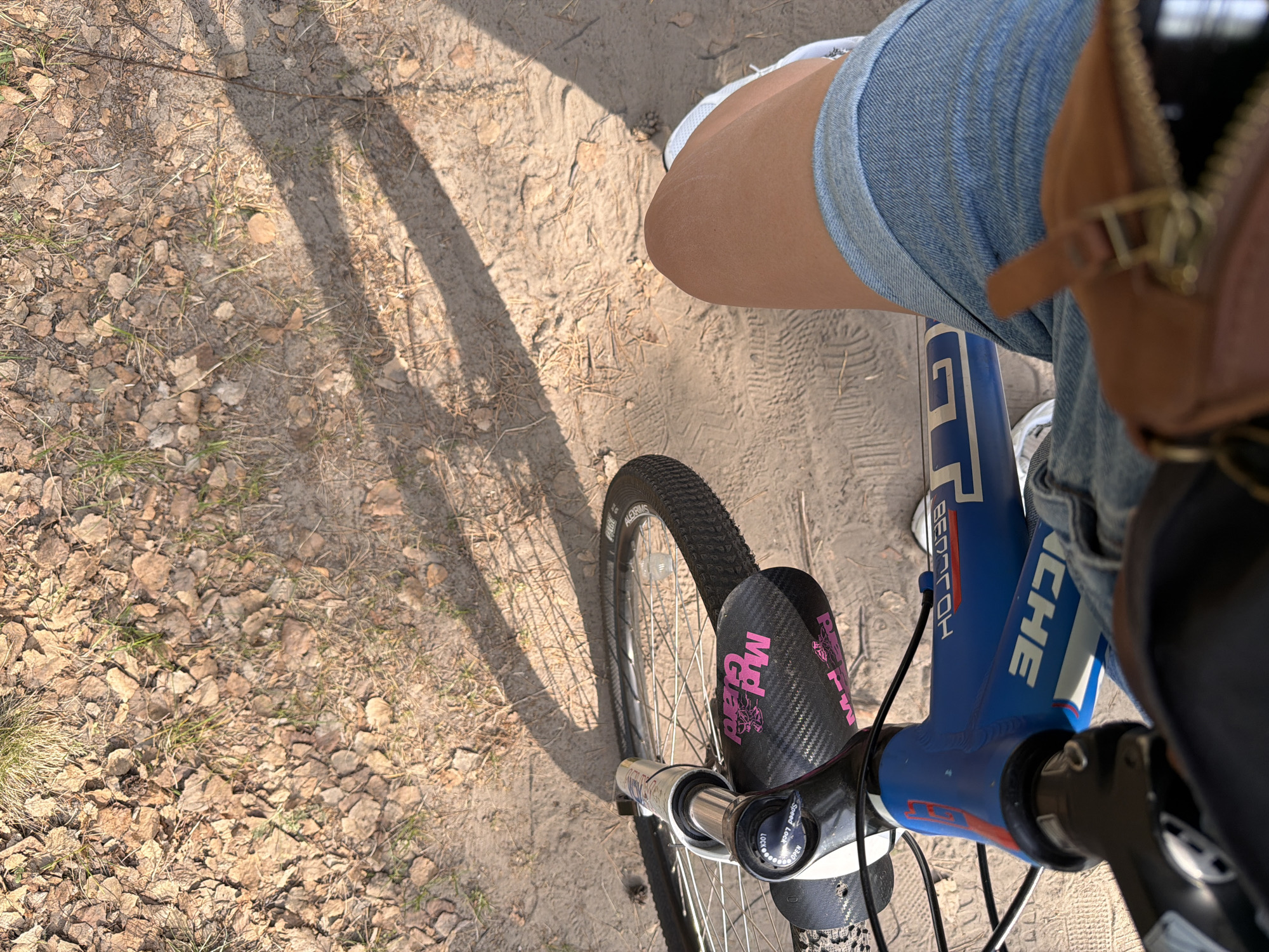 A first-person, high-angle shot depicts a person riding a bicycle on a dirt path. The path is covered in dry leaves and sparse grass. The bicycle is a blue GT model with a black front tire and a carbon fiber mudguard adorned with pink "Mtp Guard" lettering. The rider is wearing light blue denim shorts, a white shoe and a brown bag. The location cannot be ascertained to be Tyumen, Russia.