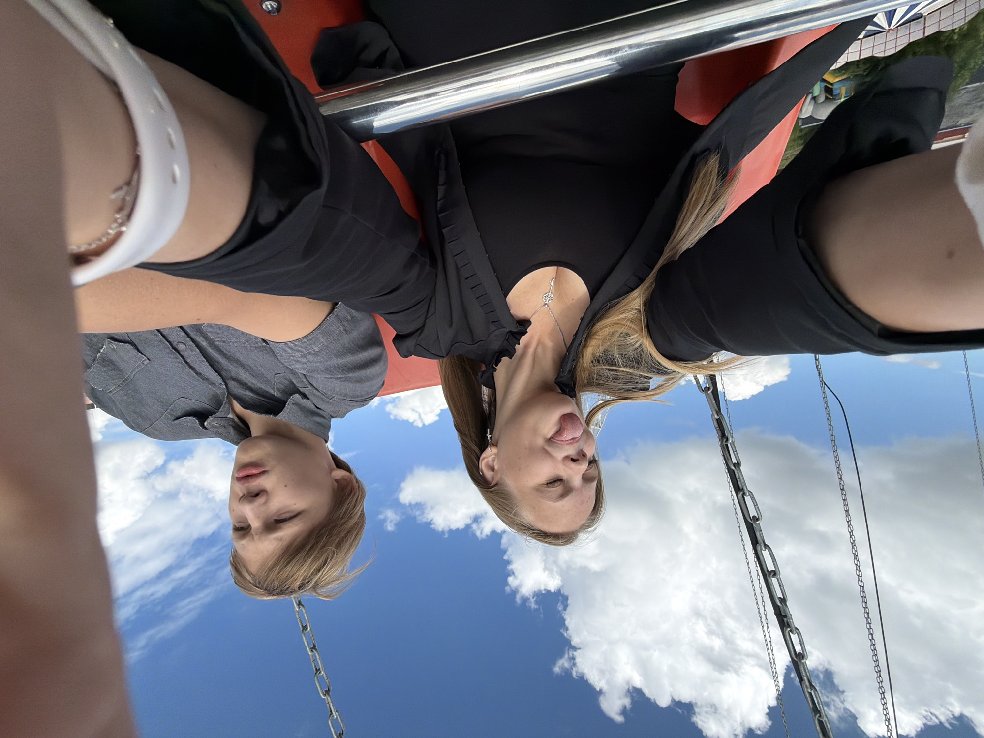 Two young people, a boy and a girl, are seen in a selfie from a first-person perspective, appearing to be on a swing ride at an amusement park. The sky is a clear blue with fluffy white clouds, suggesting it is daytime and fair weather. The photo is taken from a high vantage point, looking down at the subjects and the sky.
The boy, on the left, is wearing a grey button-up shirt. He has light brown hair and a neutral expression, looking slightly to the side.
The girl, on the right, is wearing a black top and a black jacket with ruffled details. She has long blonde hair and is sticking her tongue out with her eyes closed, conveying a playful and mischievous emotion. She is wearing a silver necklace with a key pendant. A white bracelet is visible on her arm.
The setting includes parts of the swing ride, specifically metal chains and orange seating. In the background, out of focus, are colorful structures that appear to be part of amusement park attractions. The overall impression is one of fun and lightheartedness at an outdoor entertainment venue. The image is oriented sideways.