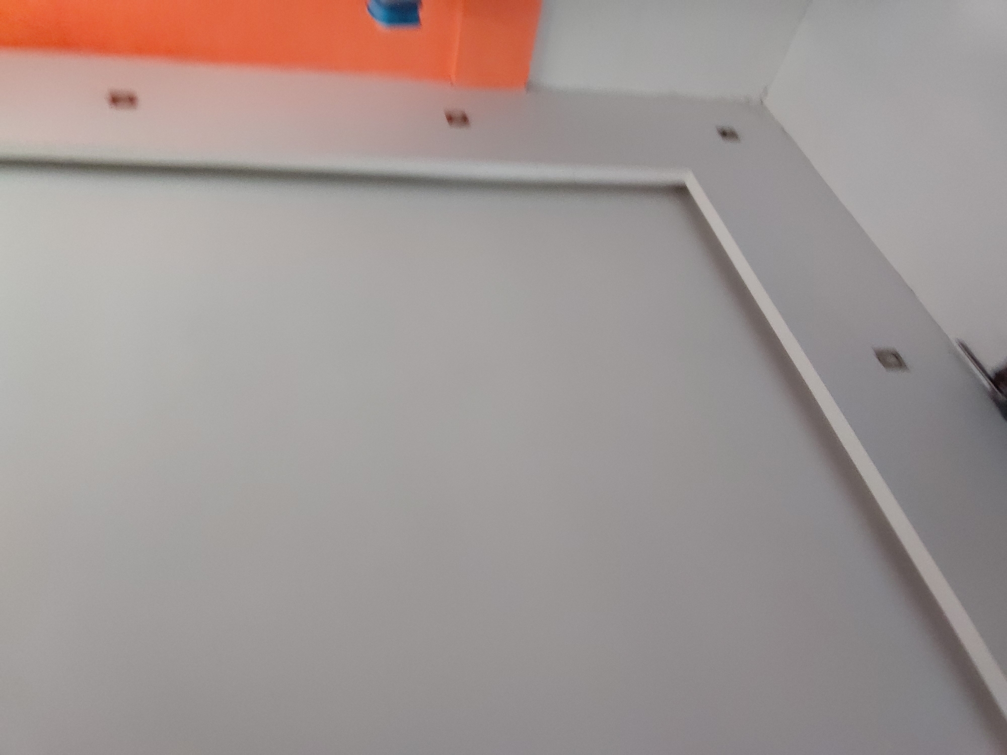 The image captures an indoor scene in Santa Ana, Venezuela, focusing on a wall and ceiling. The dominant feature is a large, light gray wall occupying the majority of the frame. To the right, a vertical section of the wall, also gray, is bordered by a crisp white frame-like structure.
The ceiling is visible at the top of the image, primarily white, but featuring a prominent bright orange section that extends across the upper left. A small, indistinct blue detail is visible on this orange ceiling segment. Several small, dark, rectangular elements, possibly electrical outlets or recessed light fixtures, are intermittently visible on both the white ceiling and the gray vertical wall section.
No people are present in the image, and no specific activity is occurring. The scene appears static, possibly depicting a room during or after construction or renovation. The lighting is even, providing no cues about the time of day or external weather. There is no visible text within the image. The overall impression is of a clean, somewhat minimalist interior space.