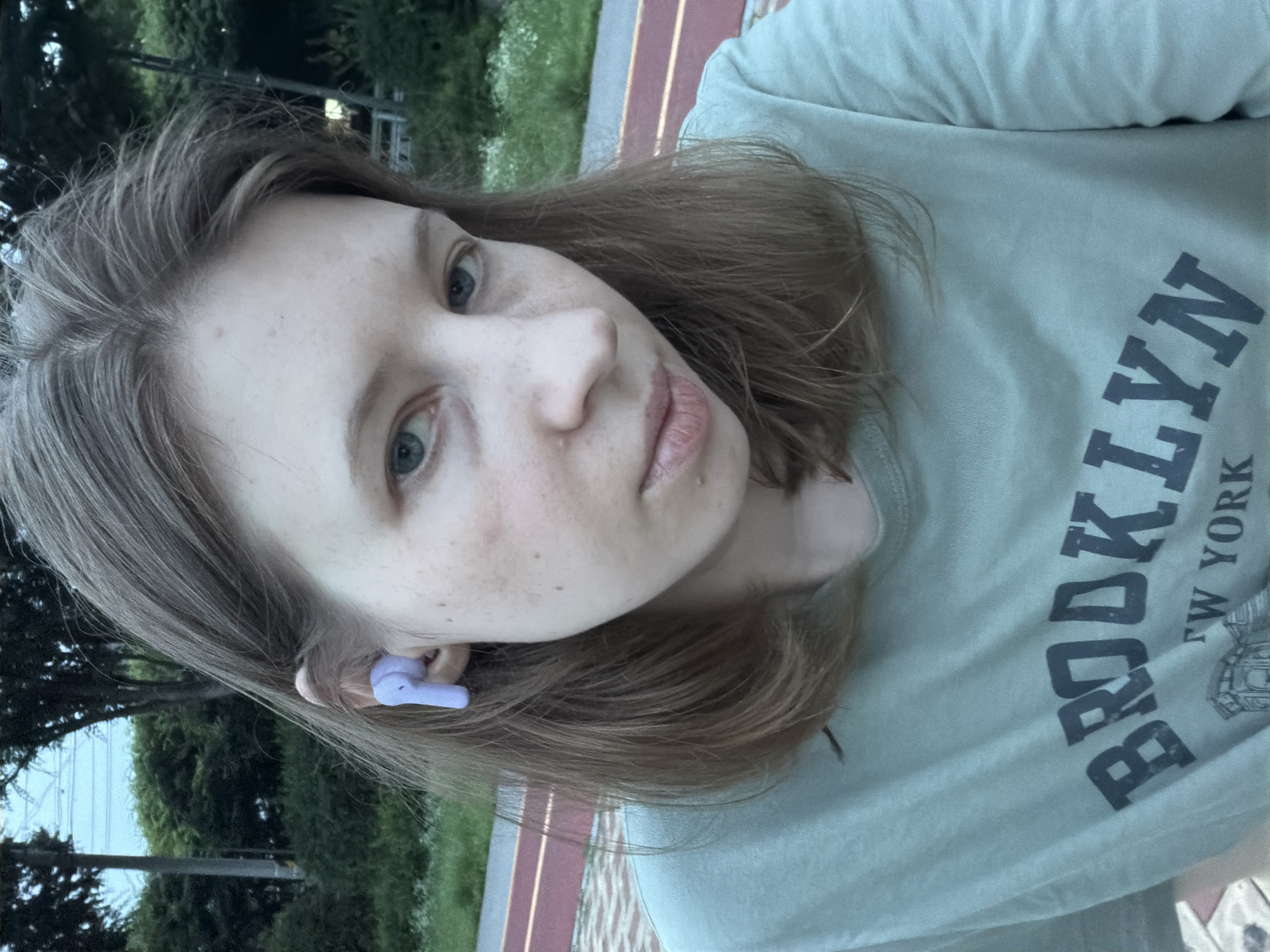 A young woman with shoulder-length brown hair and blue eyes is captured in a close-up, outdoor selfie. She is wearing a light green t-shirt with the text "BROOKLYN NEW YORK" printed on it. A light purple earbud is visible in her right ear. The background features lush green trees and foliage, suggesting a park or natural setting. The ground is paved with a reddish track and lighter paving stones. The overall lighting indicates it is daytime, possibly late afternoon or early evening. The location is the city of Dongducheon-si, South Korea.