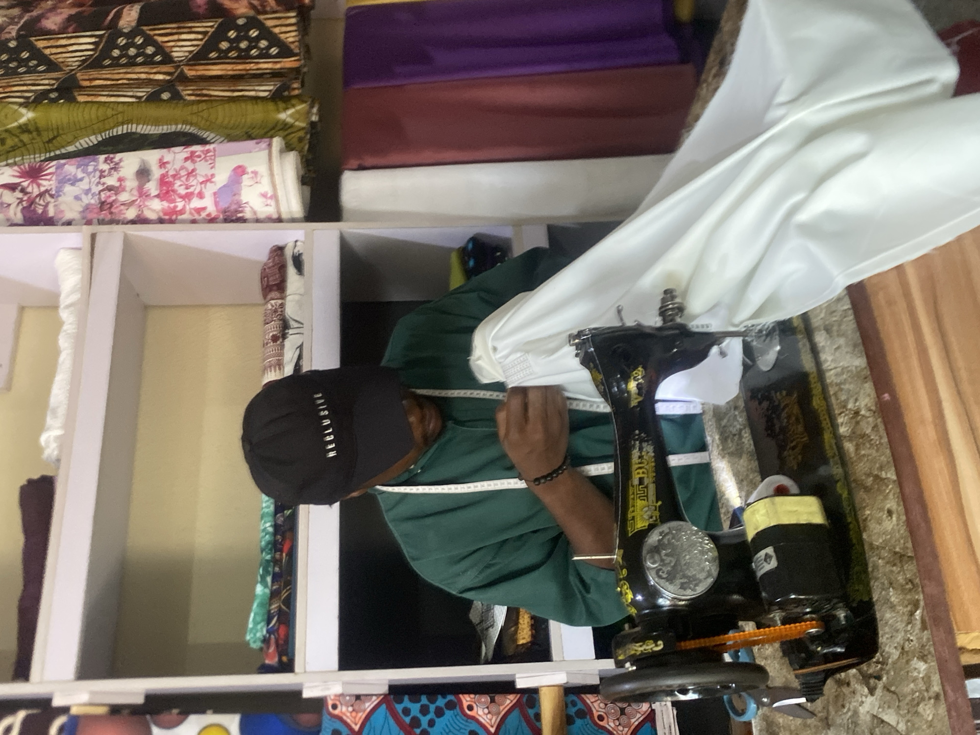 In a tailor's workshop in Bukuru, Nigeria, a man wearing a green shirt and a black cap with "RECLUSIVE" printed on it is operating a vintage black sewing machine. He is focused on sewing a white fabric, holding it in place with his hands. A tape measure is draped around his neck. The workshop is filled with bolts of colorful fabrics, stacked on shelves and hanging on a rack. The shelves display various textiles, including patterned cloths and solid colored materials. The sewing machine itself has a visible motor and a prominent handwheel. The lighting suggests an indoor setting, likely during the day. The scene depicts a typical small business environment focused on garment production.